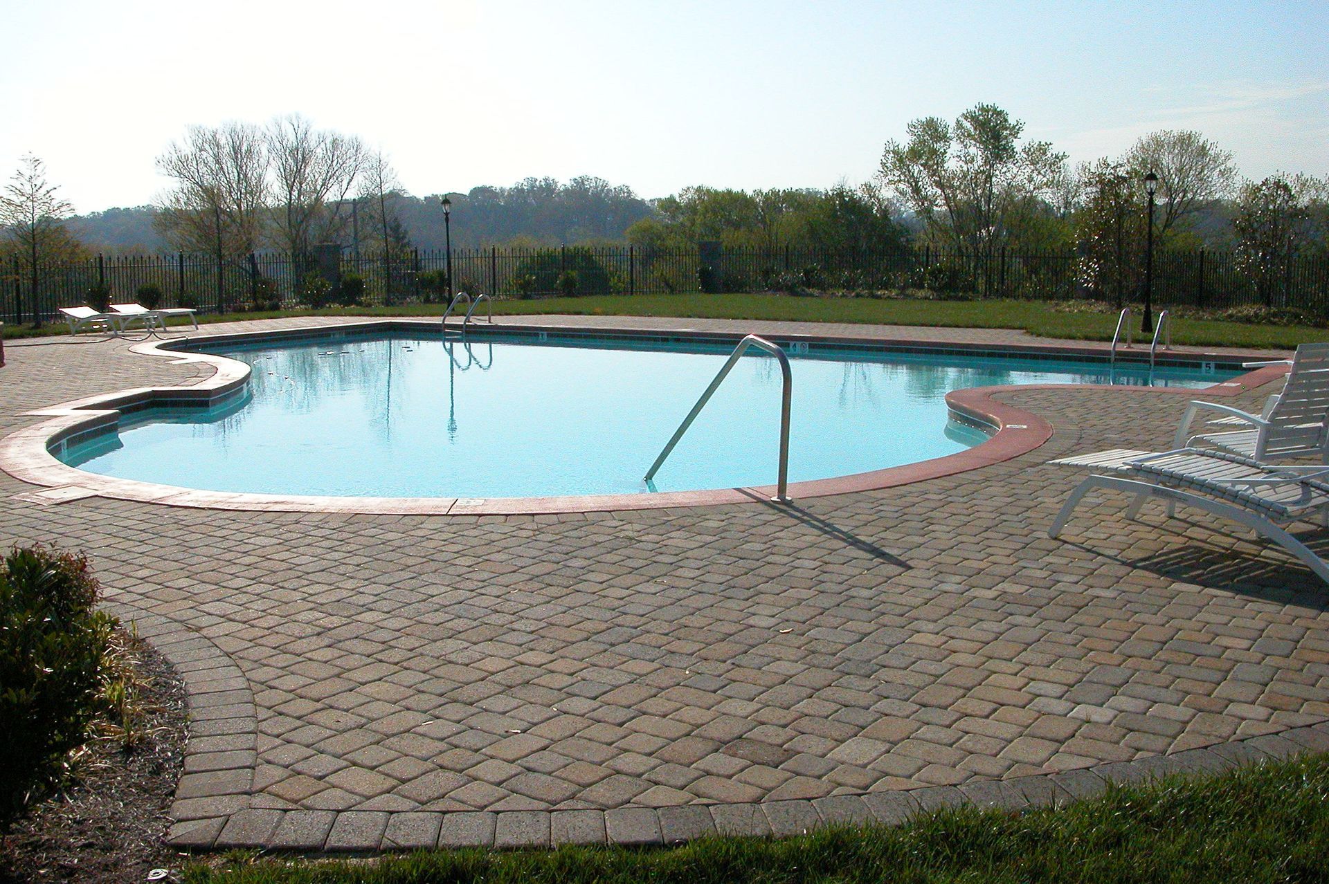 Outdoor swimming pool with curved blue water, brick deck, lounge chairs, and grassy trees in the background