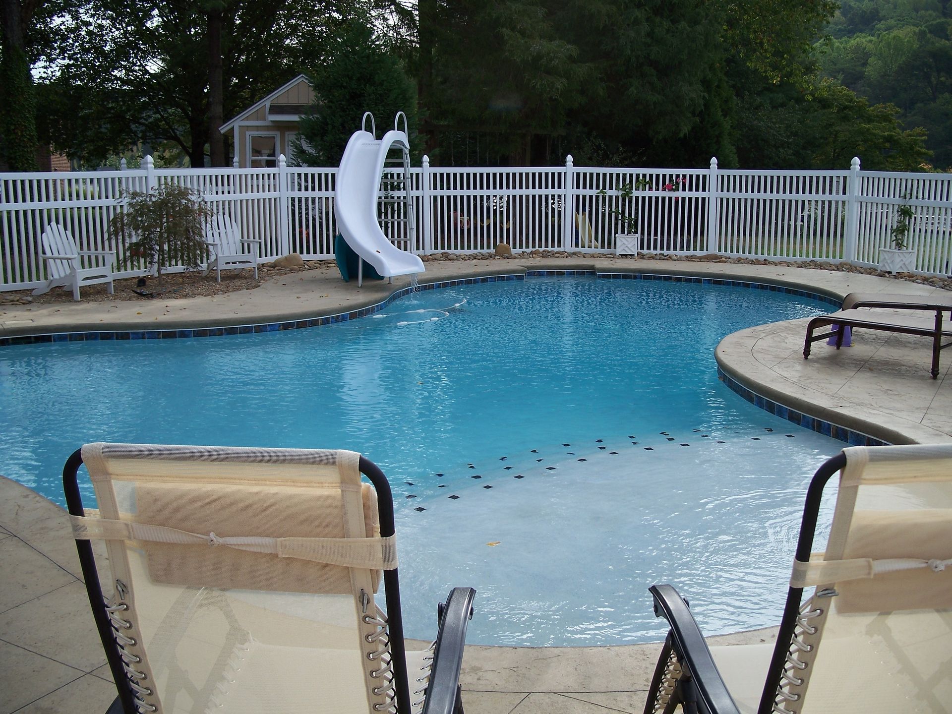 Backyard swimming pool with white fence, curved slide, and lounge chairs in the foreground