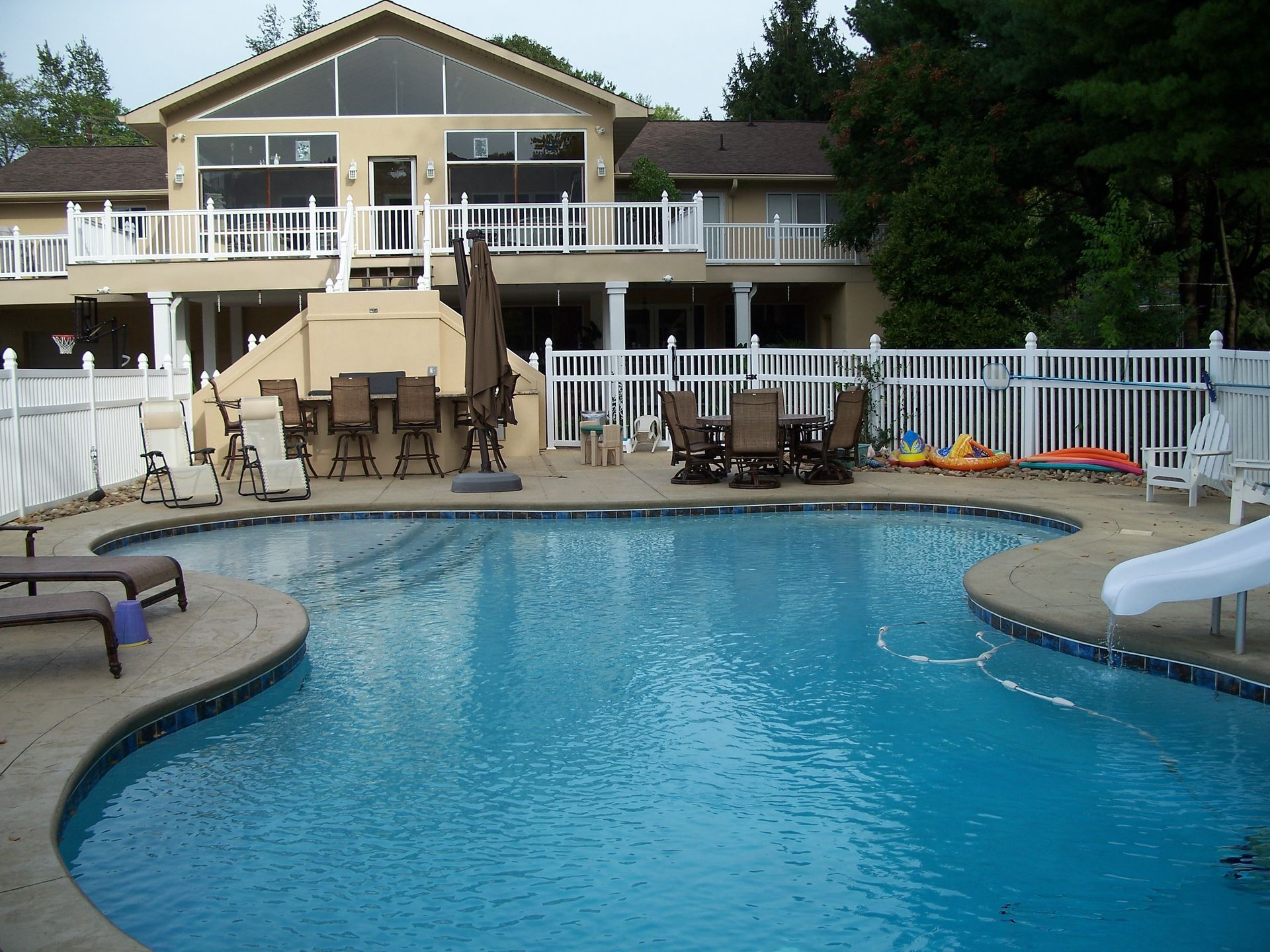 Backyard pool with patio, white fence, lounge chairs, and a house in the background