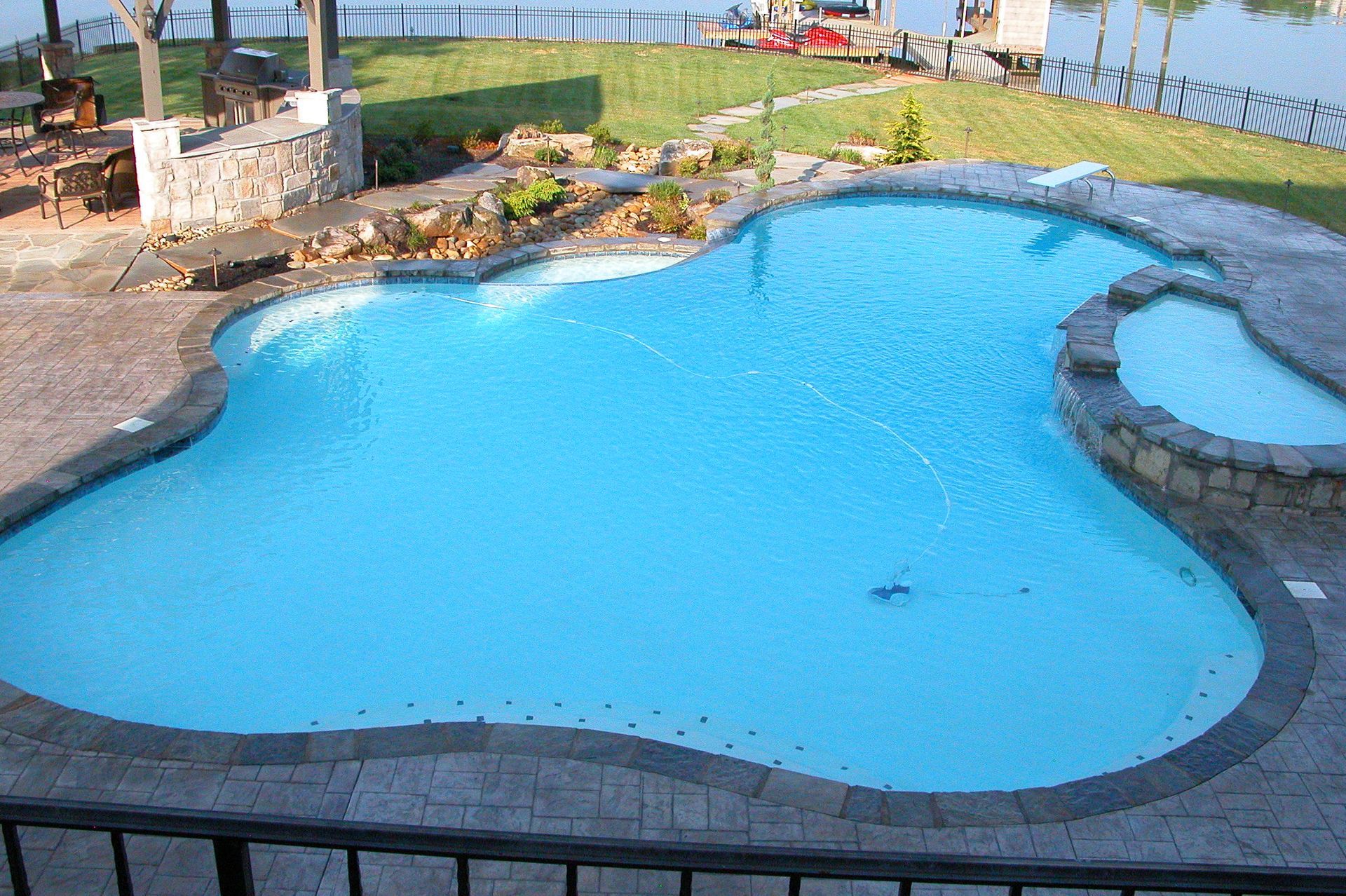 Curved blue swimming pool with stone edging and a small raised spa area in a grassy backyard