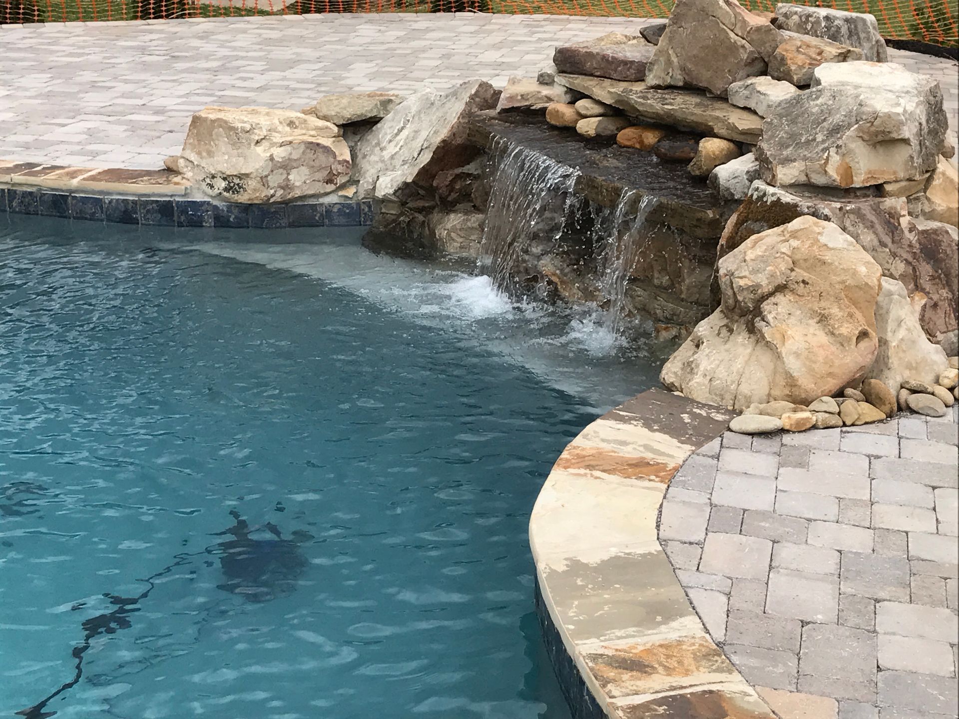Backyard swimming pool with a stone waterfall and blue water beside a paver patio