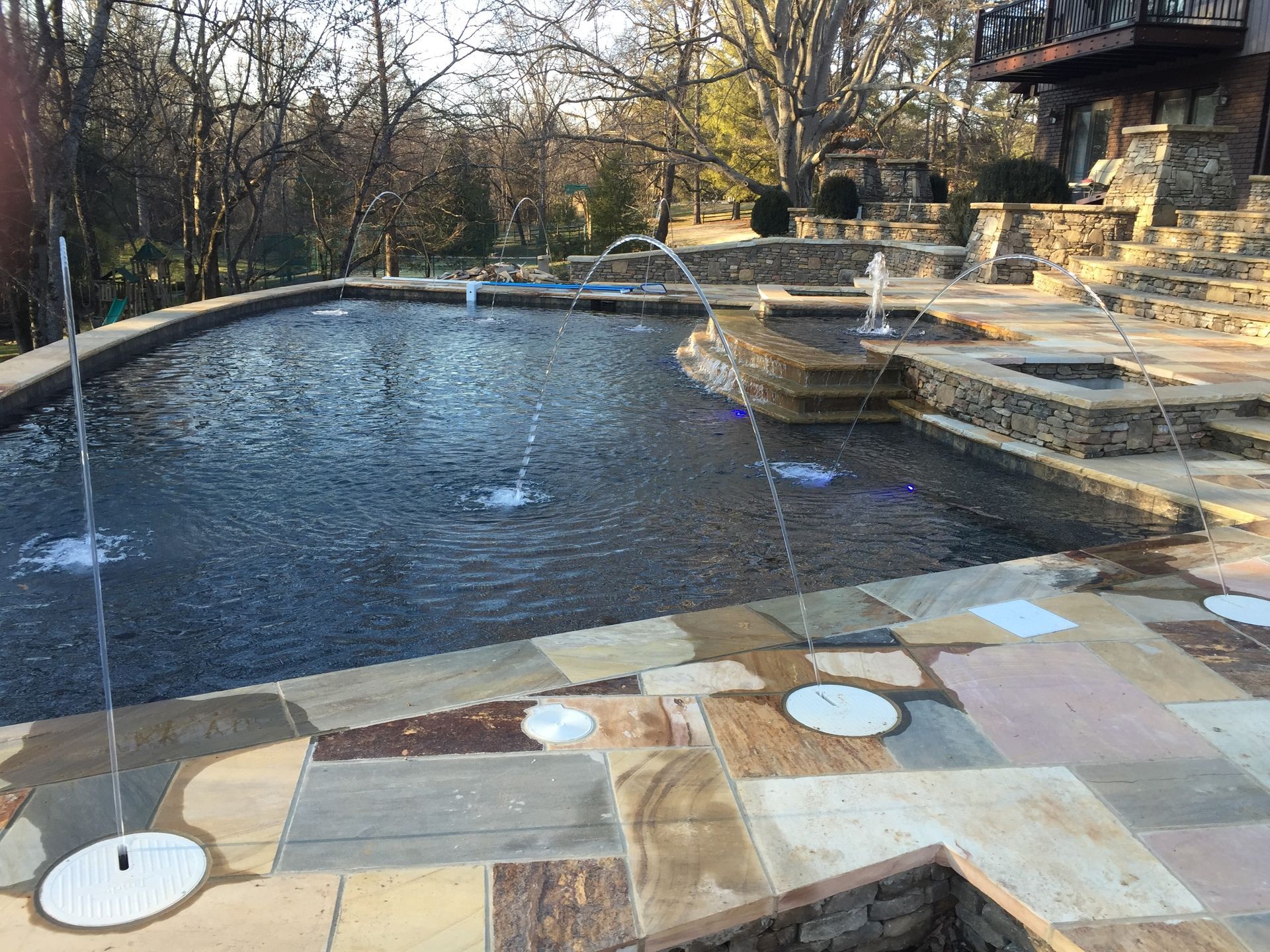 Curved backyard pool with fountains, stone patio, and a house in the background
