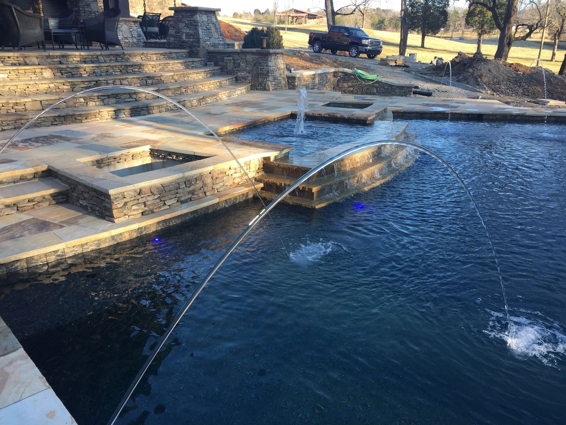 Garden pond with a stone fountain spraying water into a dark blue pool, bordered by steps and landscaping.