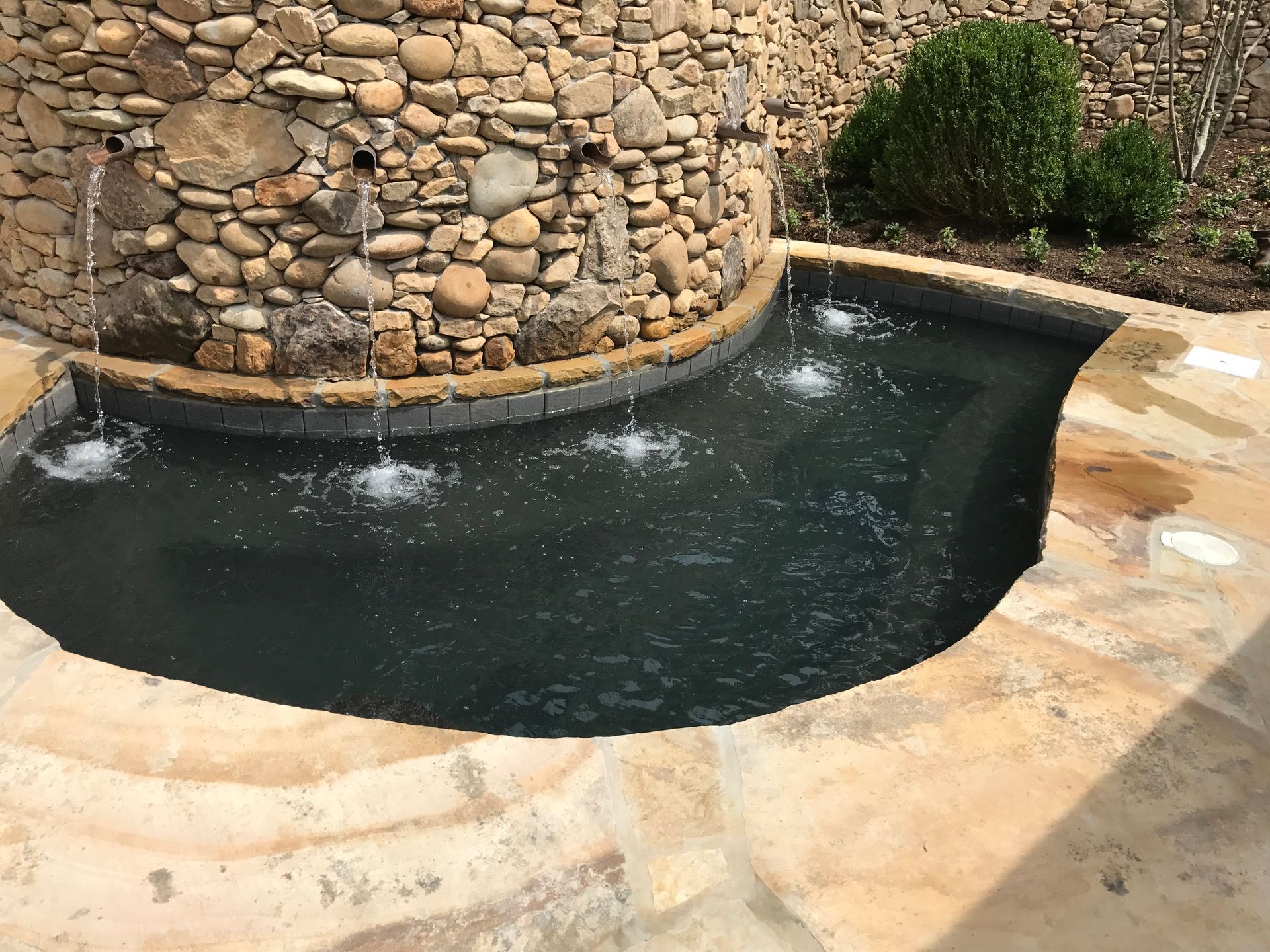 Small stone-lined backyard pool with dark water and two bubbling fountains beside a tan patio.