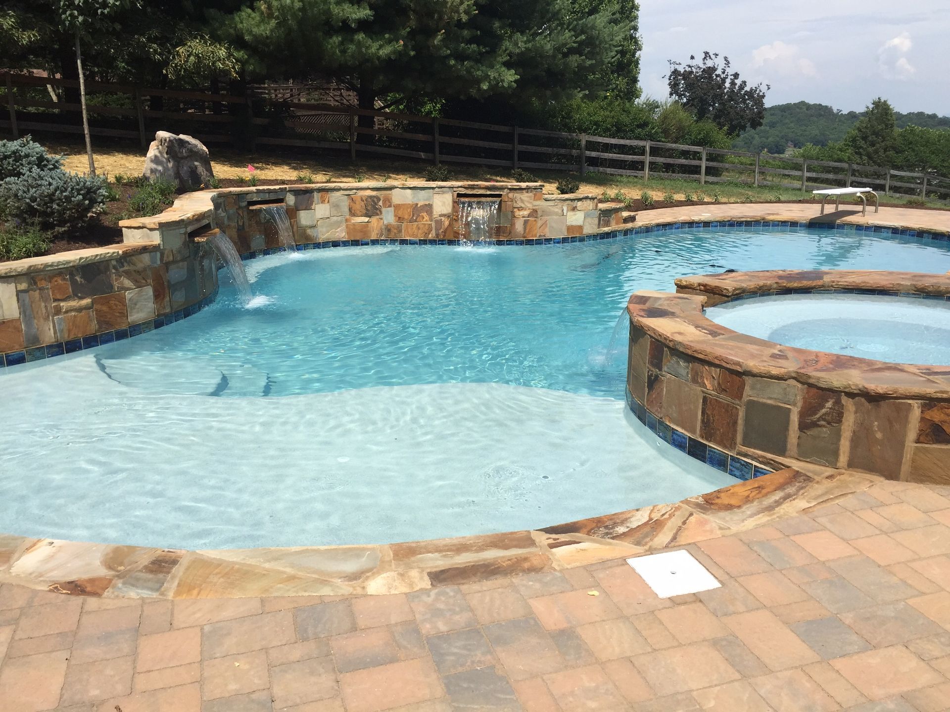 Backyard pool with a curved blue swimming area, stone edging, and a round hot tub on a brick patio.
