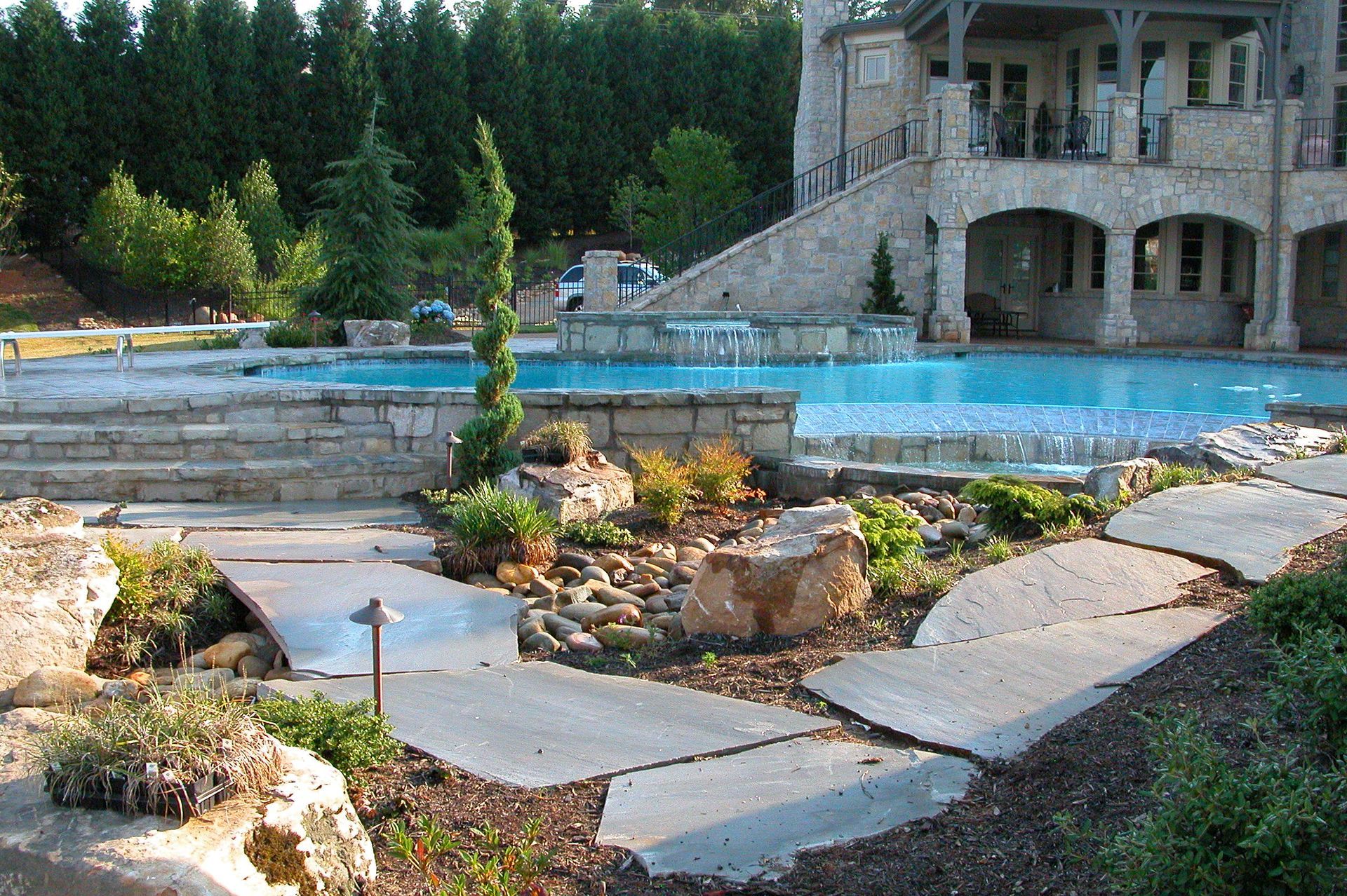 Stone garden walkway around a blue pool beside a house and stone staircase