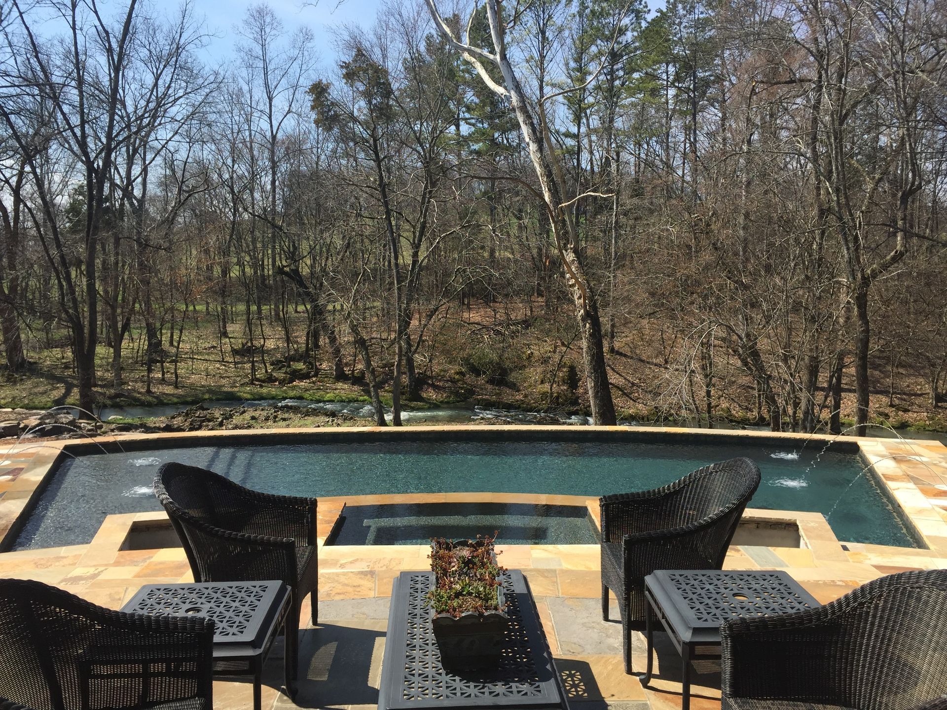 Patio chairs around a fire pit beside a swimming pool with wooded trees in the background