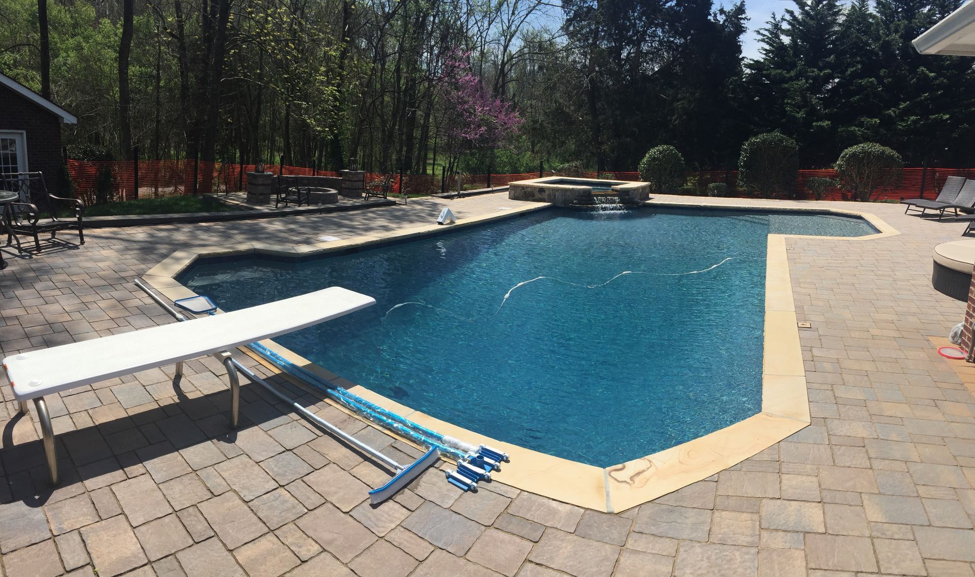 Outdoor swimming pool with a diving board, surrounded by paving and trees on a sunny day