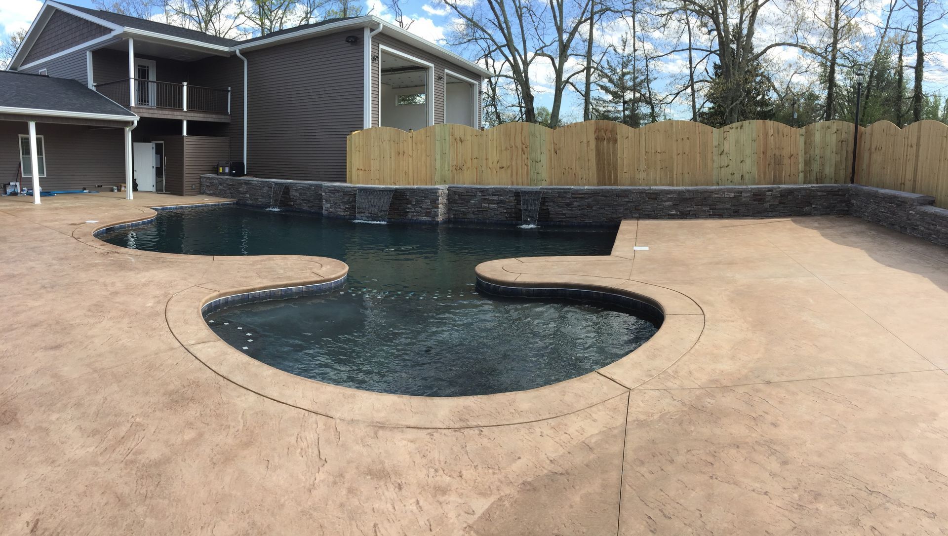 Backyard swimming pool with irregular shape and concrete deck beside a house and wooden fence