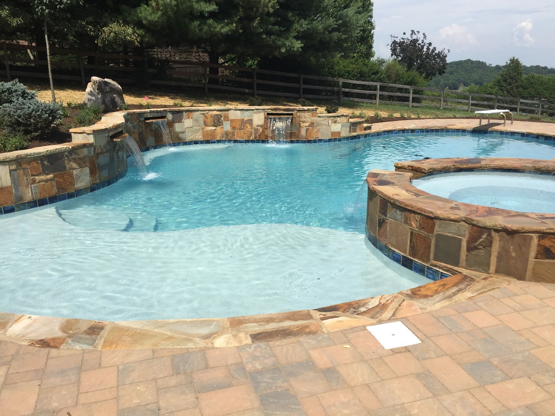 Outdoor swimming pool with curved stone edges, shallow blue water, and a small hot tub area nearby.