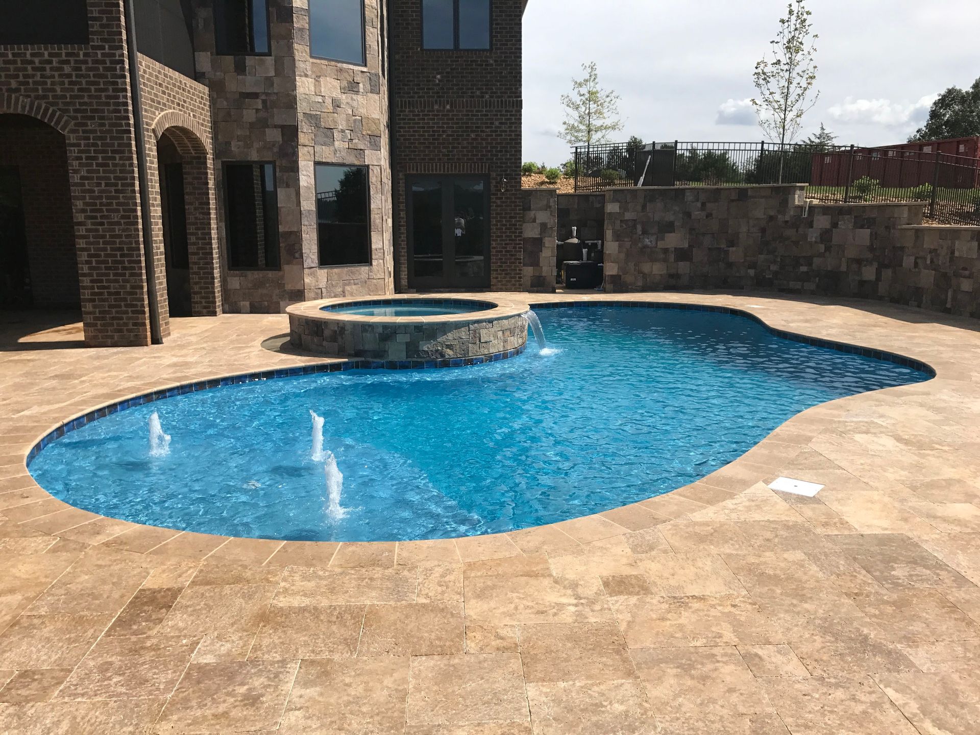 Backyard pool with blue water, tan stone deck, and a raised circular spa beside a brick house.