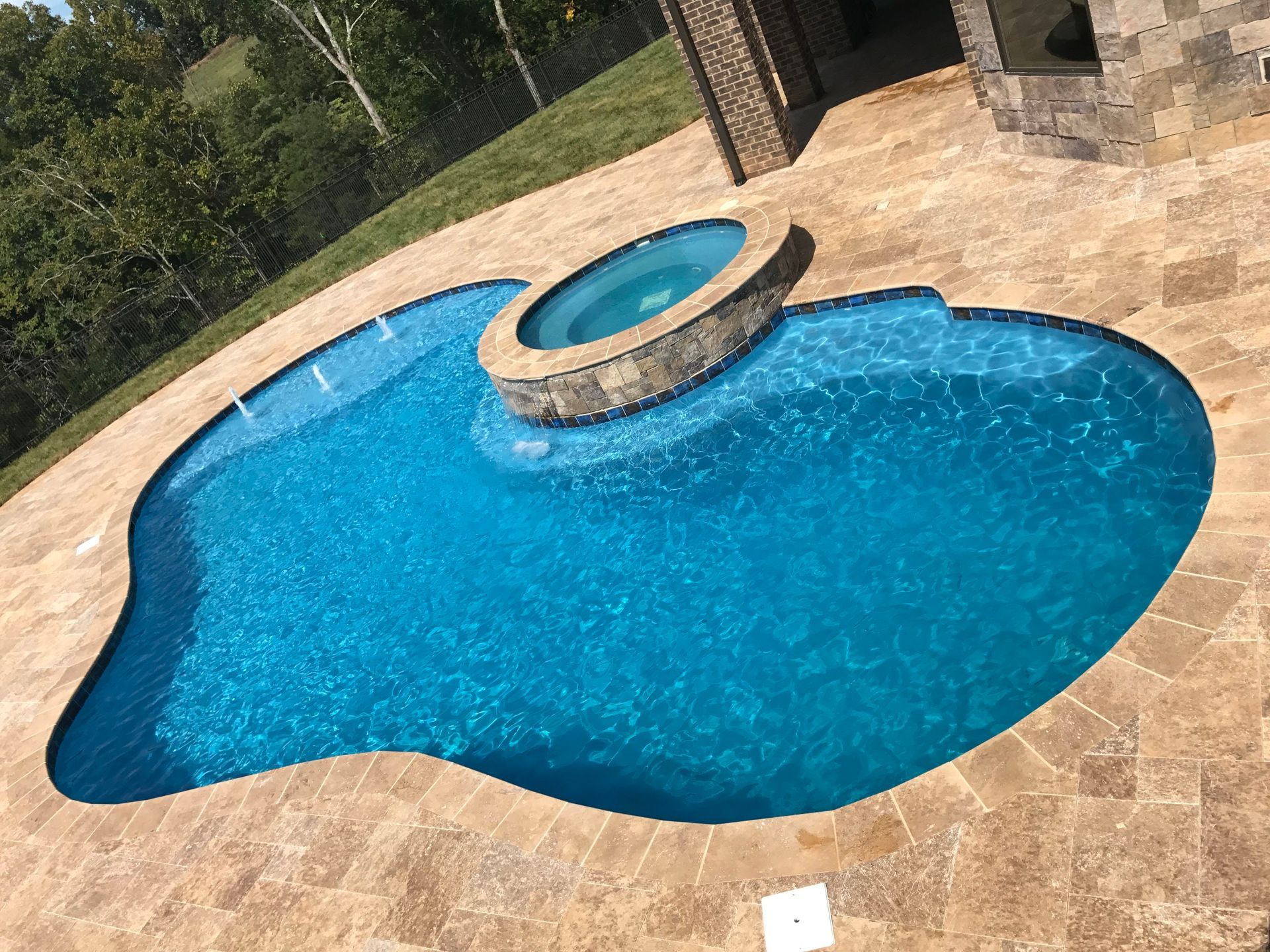 Curved blue backyard pool with tan stone deck and raised circular spa