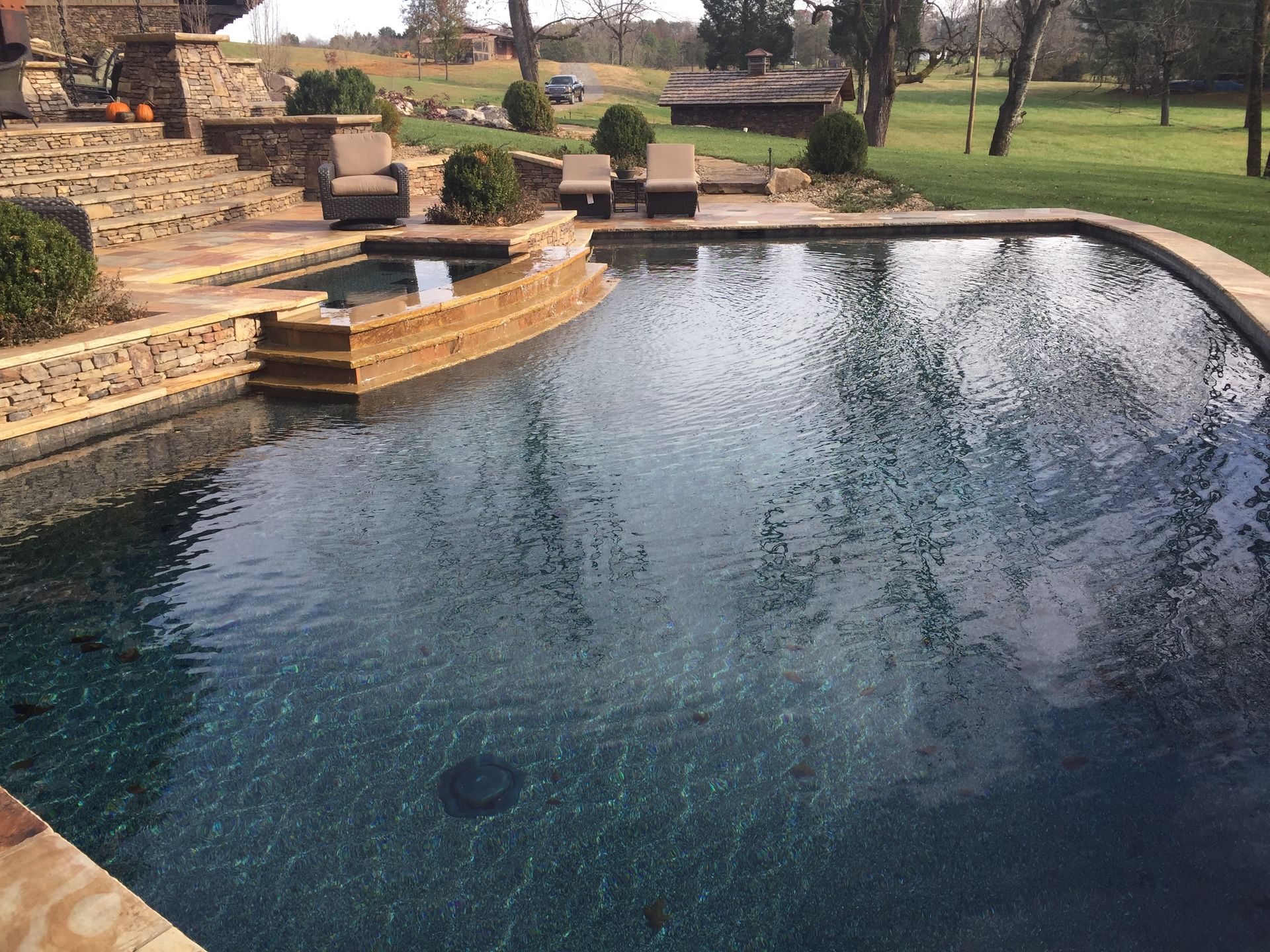 Stone-lined swimming pool with clear blue water beside a grassy park and patio seating