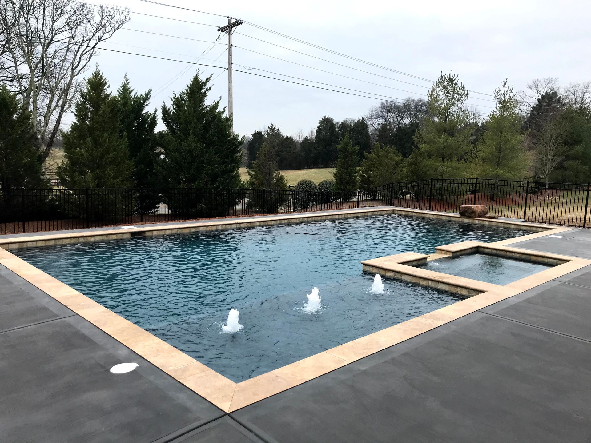 Rectangular backyard pool with four fountain jets, beige coping, and trees in the background