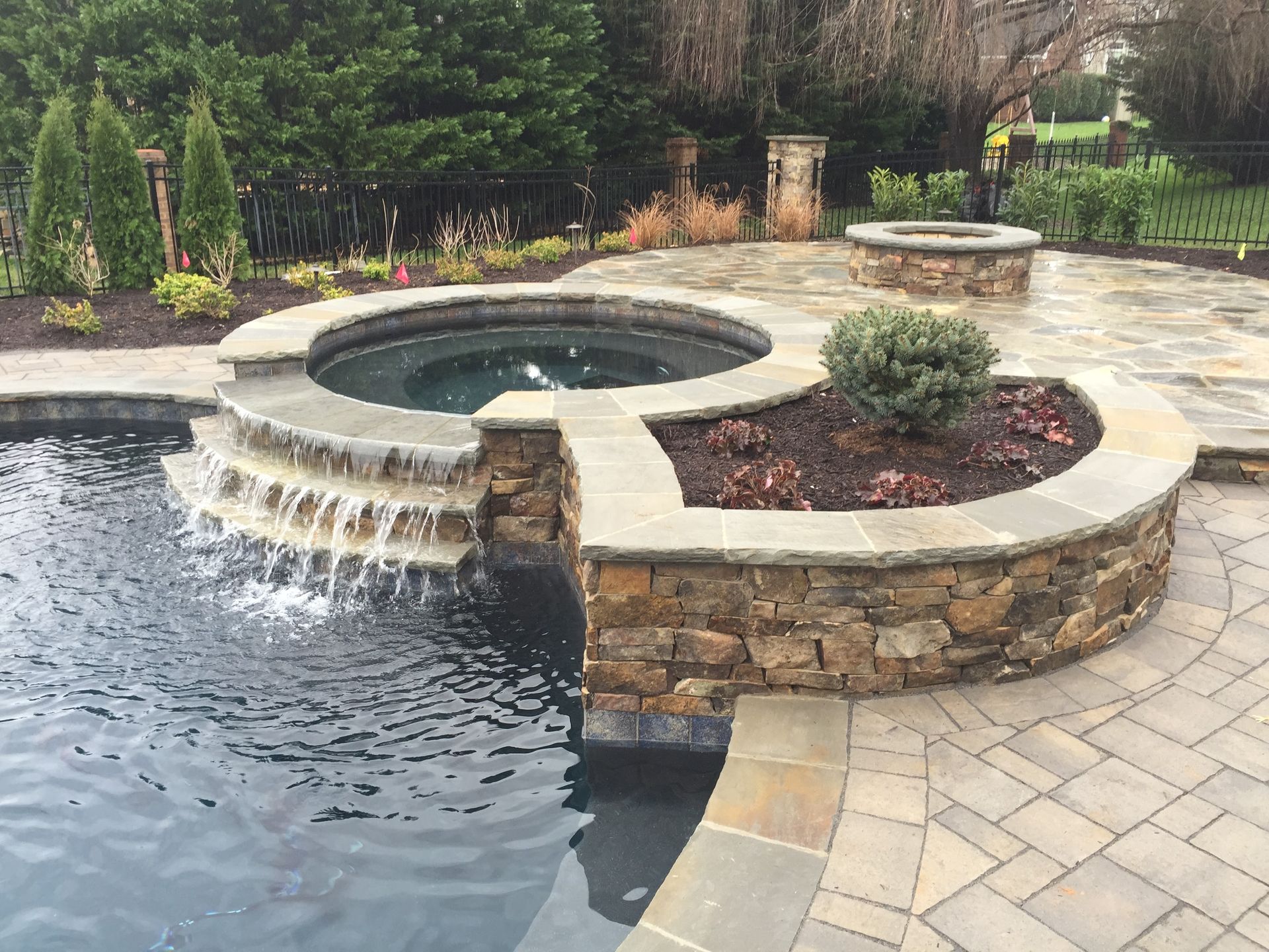 Stone hot tub with cascading water beside a landscaped patio and poolside garden.