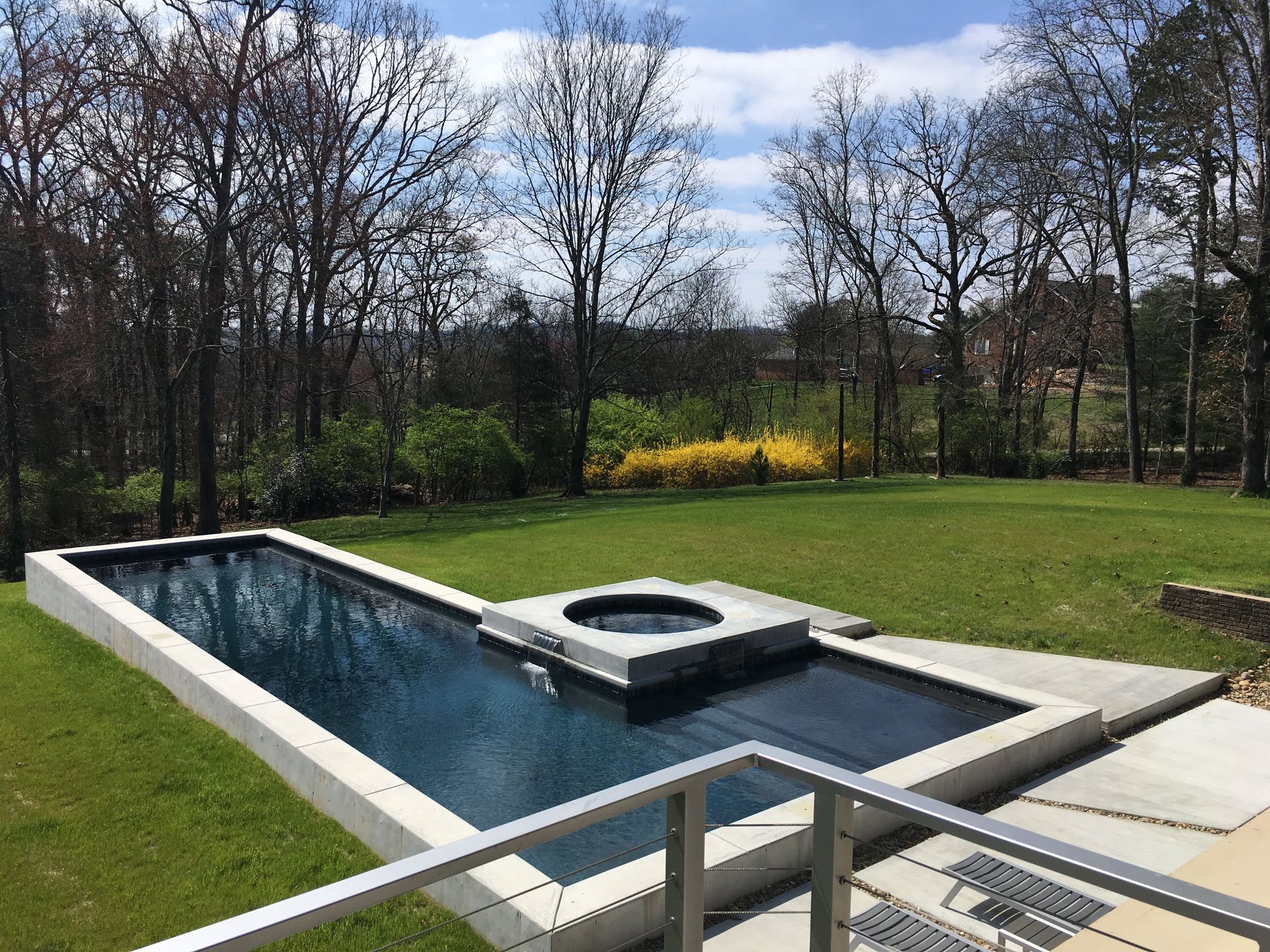 Backyard swimming pool with a hot tub, bordered by grass and trees on a cloudy day