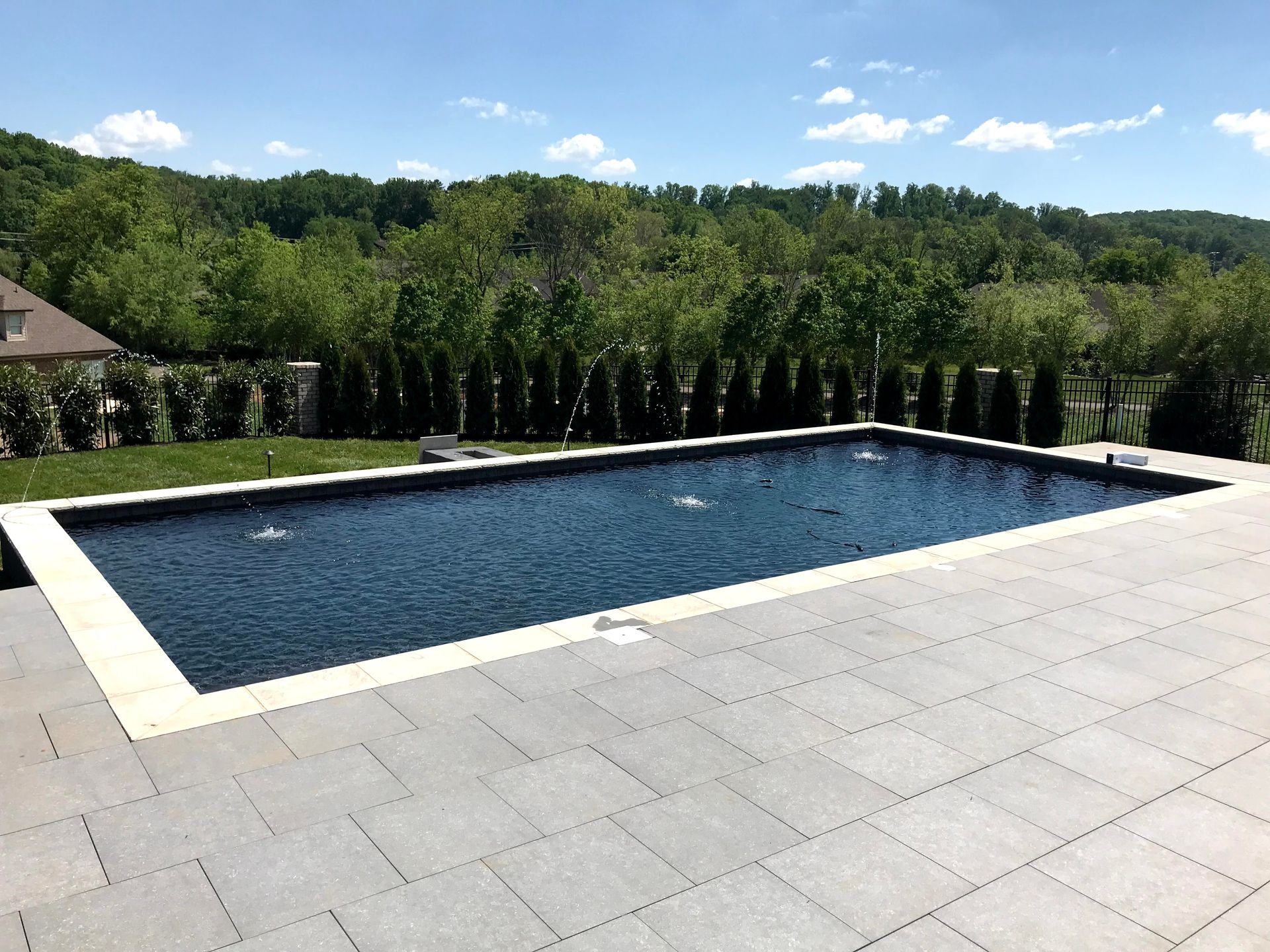 Rectangular backyard swimming pool with dark blue water, stone patio, and green trees beyond
