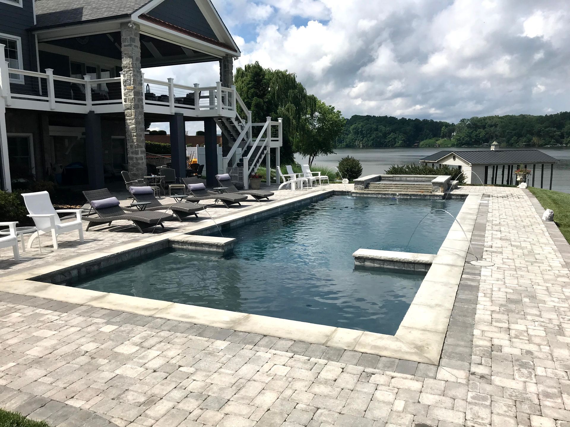 Modern lakeside pool beside a gray house, with lounge chairs, dock, and cloudy sky.