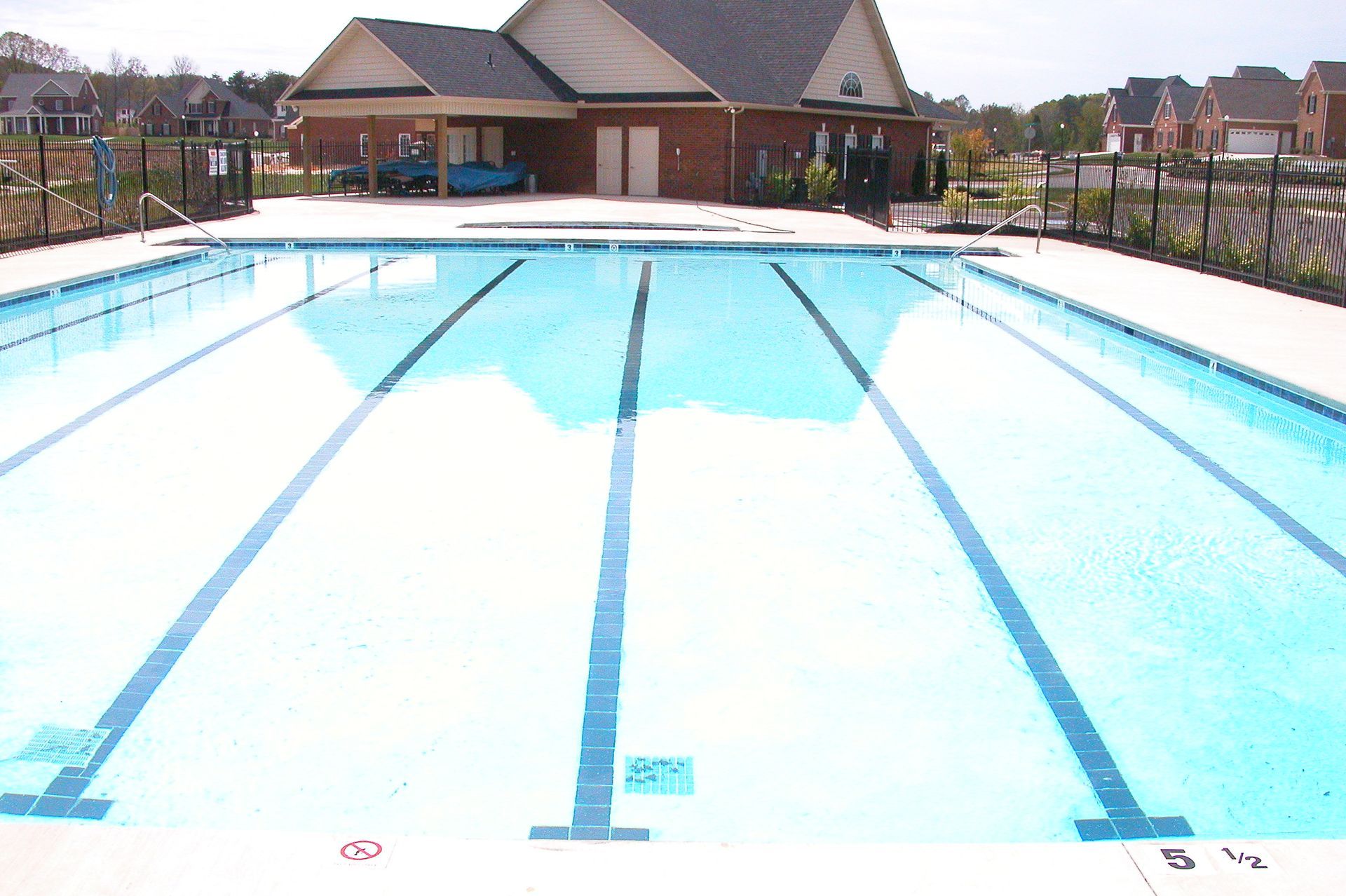 Outdoor swimming pool with lane markings under bright sun, surrounded by a residential clubhouse and houses.