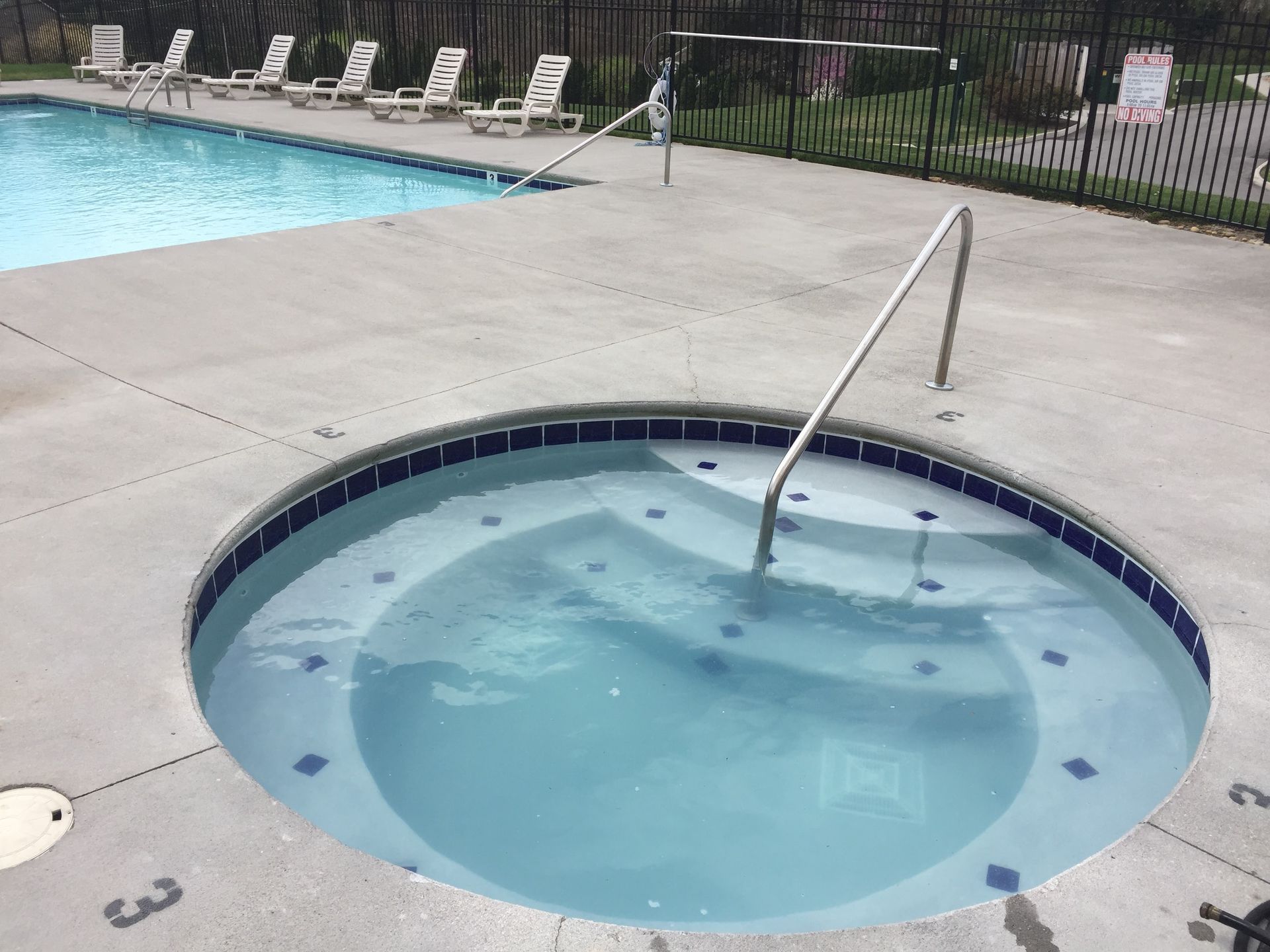 Small round hot tub beside a swimming pool, with a metal handrail and lounge chairs in the background.
