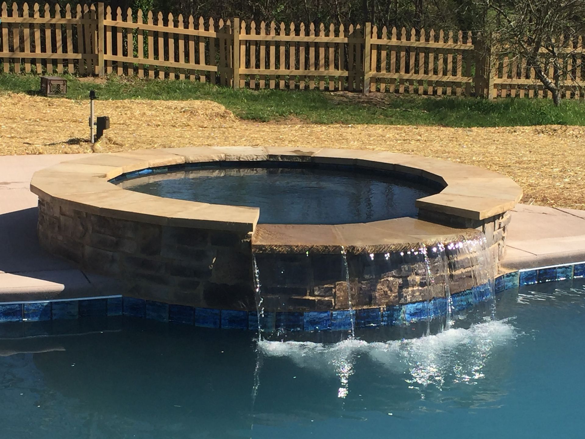 Round hot tub beside a blue pool, with a wooden fence and dry grass in the background.