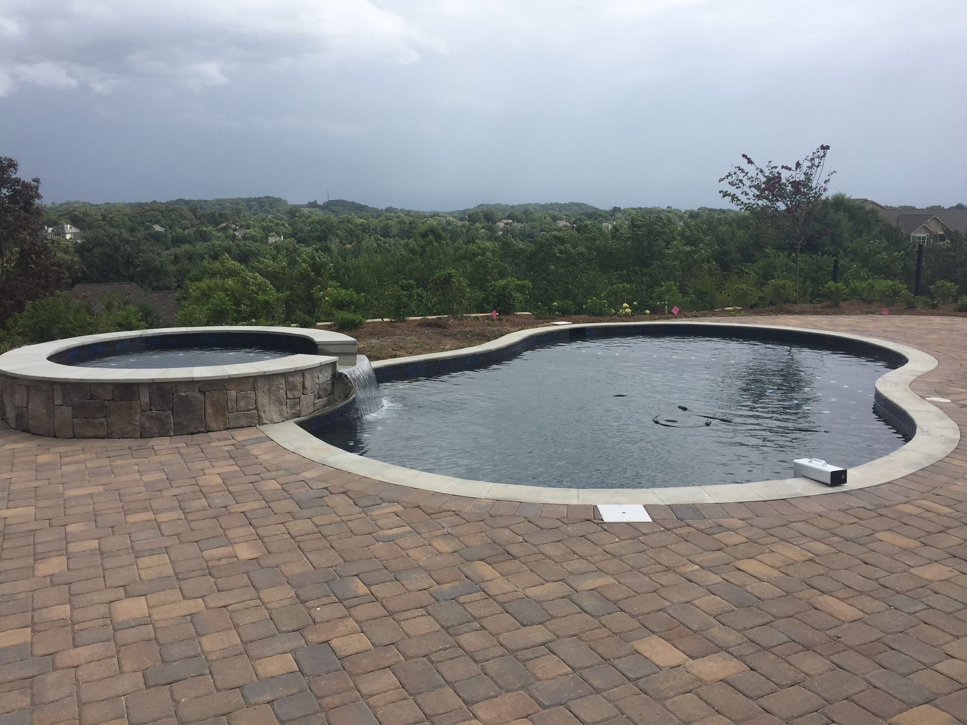 Stone pool and hot tub beside a brick patio, overlooking green trees under a cloudy sky.