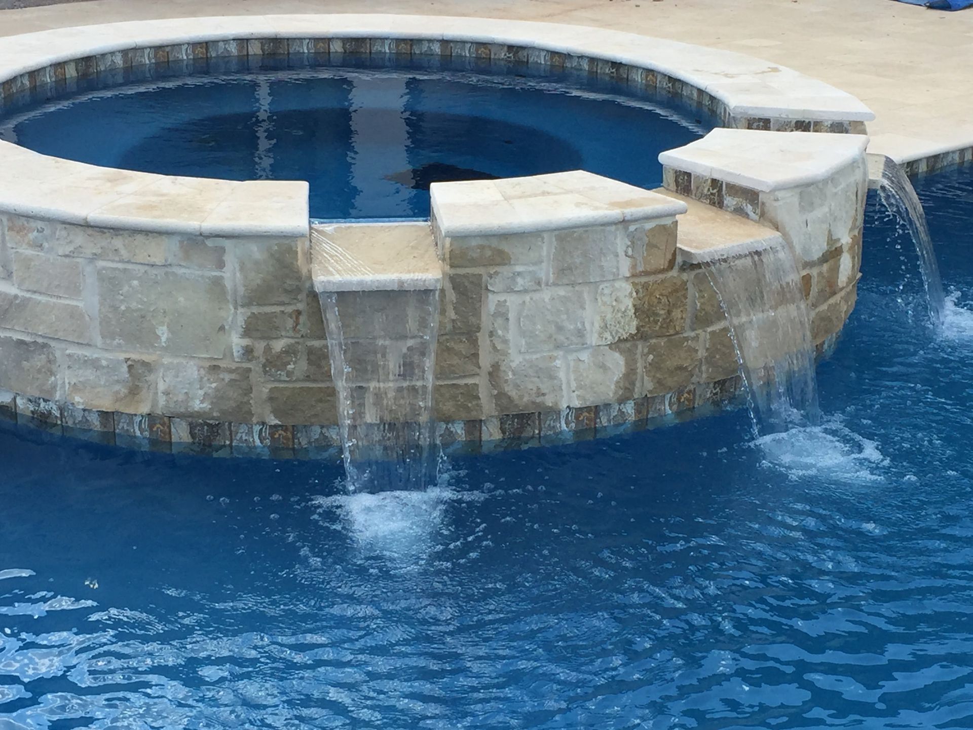 Round stone hot tub with blue water and a small waterfall spilling into a swimming pool