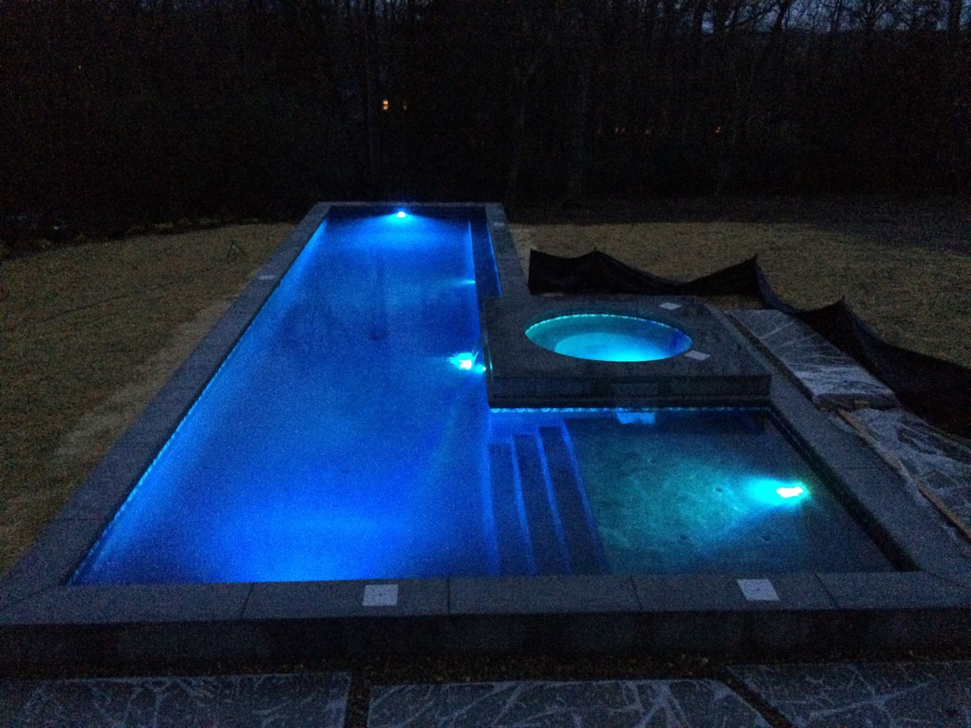 Blue-lit backyard pool at night with a hot tub and deck
