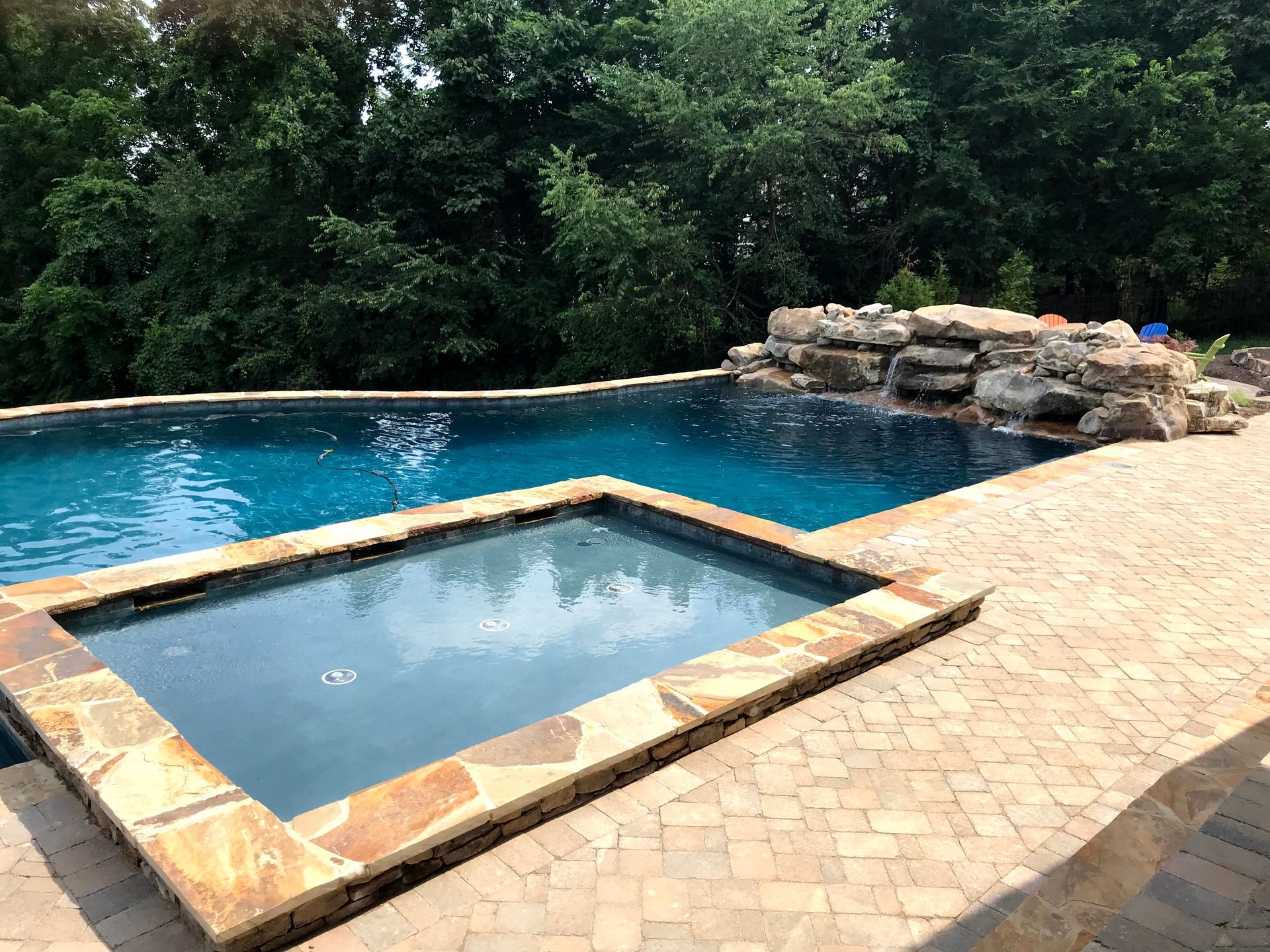 Backyard pool with a square spa, stone deck, and lush green trees in the background