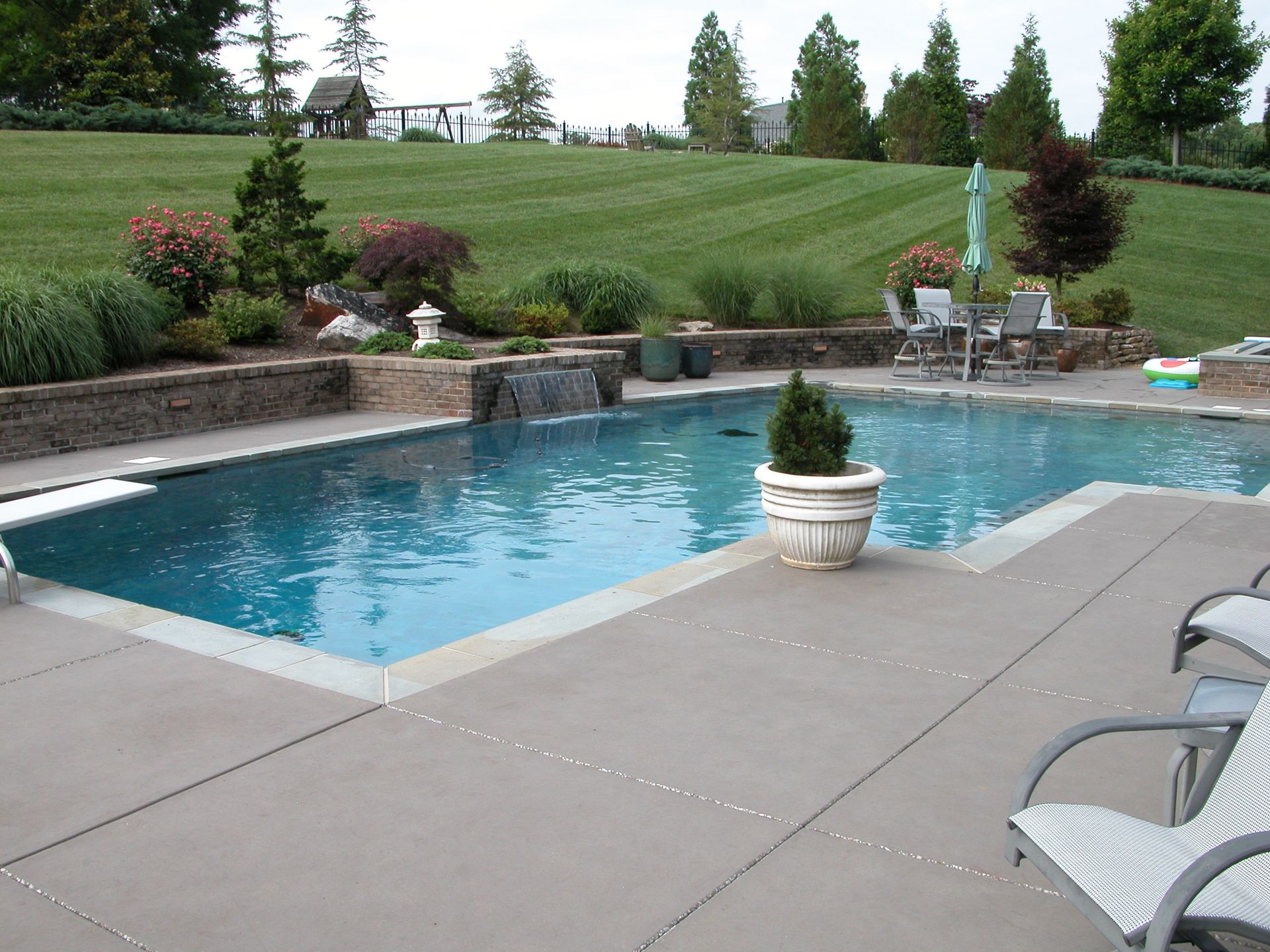 Backyard swimming pool with patio chairs, potted plants, and a manicured lawn beyond