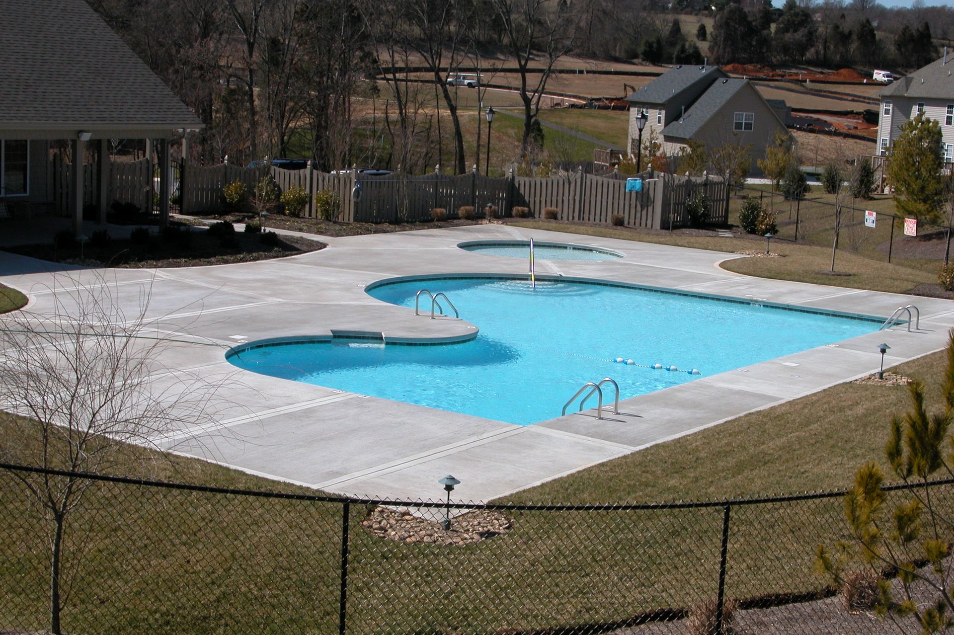 Empty outdoor swimming pool with bright blue water, surrounded by concrete and a chain-link fence.