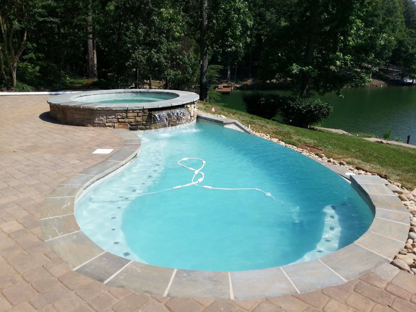 Curved turquoise hot tub beside a stone patio, with a circular raised spa and wooded lakeside view.