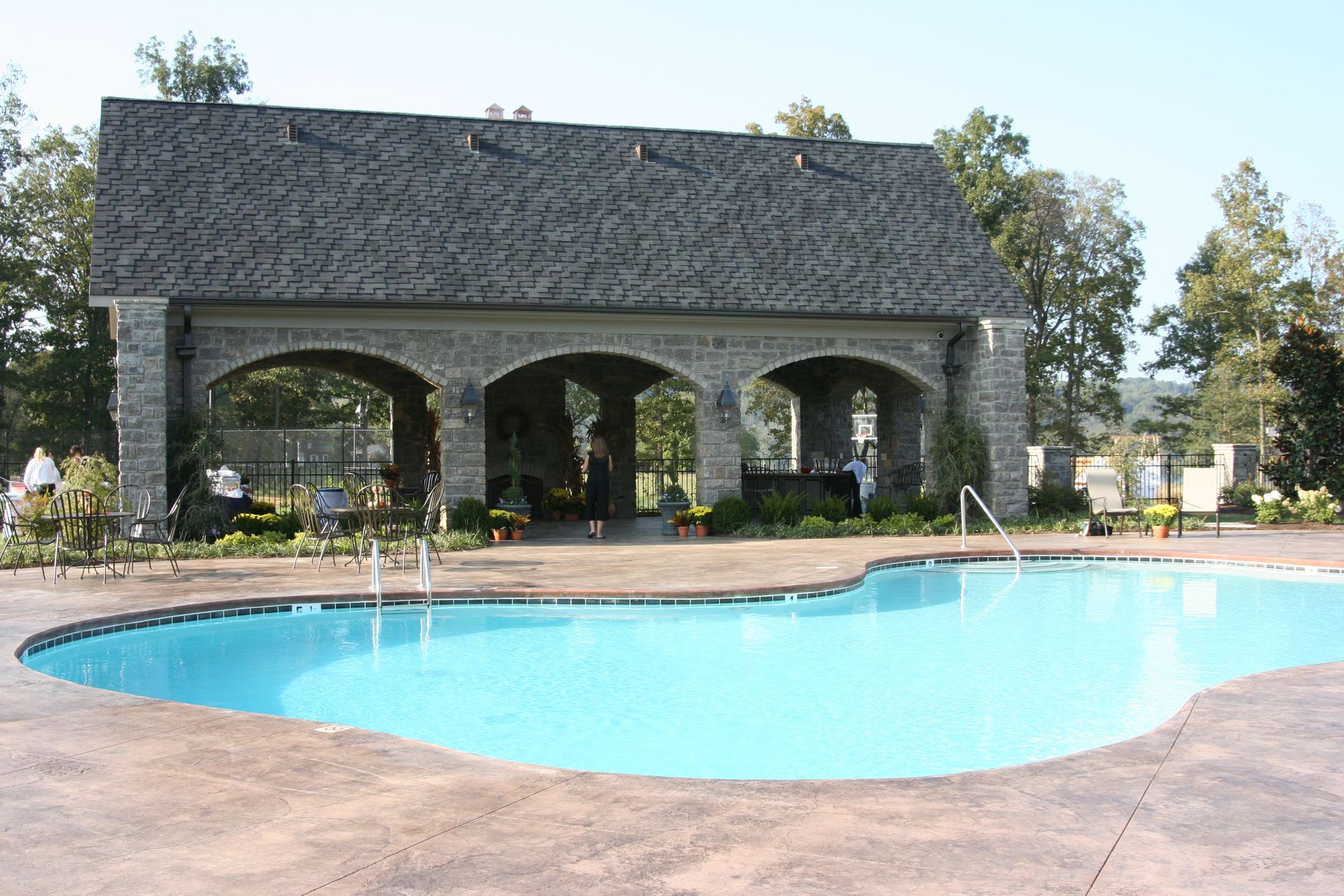 Outdoor swimming pool beside a stone pavilion with arched openings and trees in the background