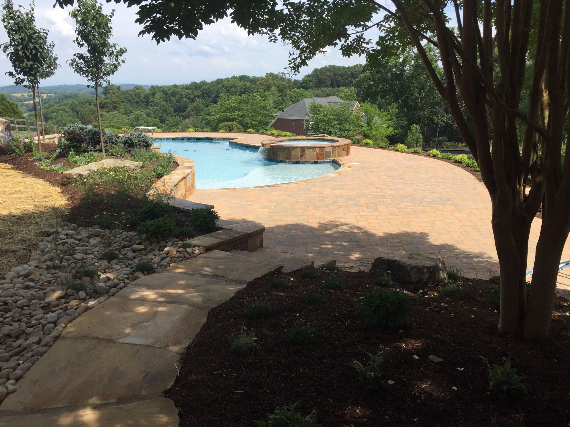 Curved pool with stone deck, trees, and landscaped garden overlooking a green hillside