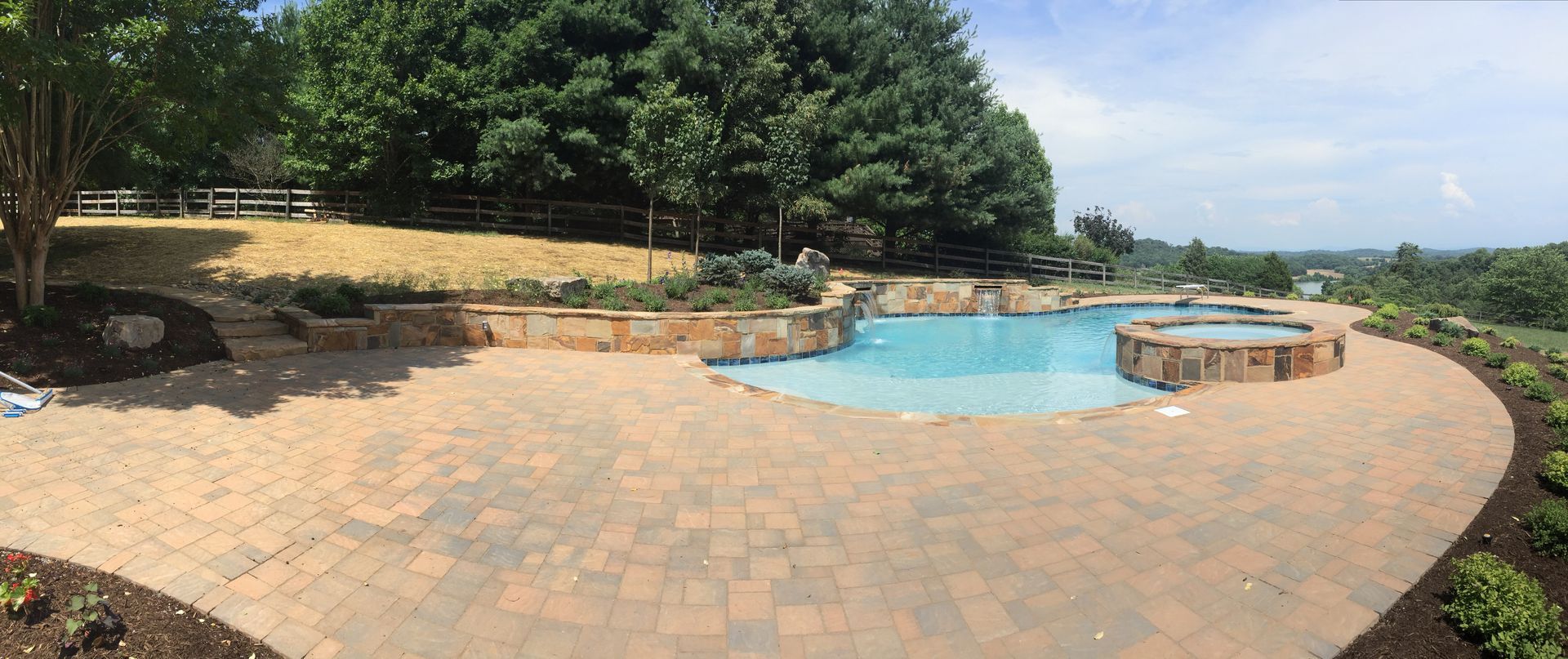 Curved brick patio with a blue pool, stone seating, and trees overlooking a distant landscape