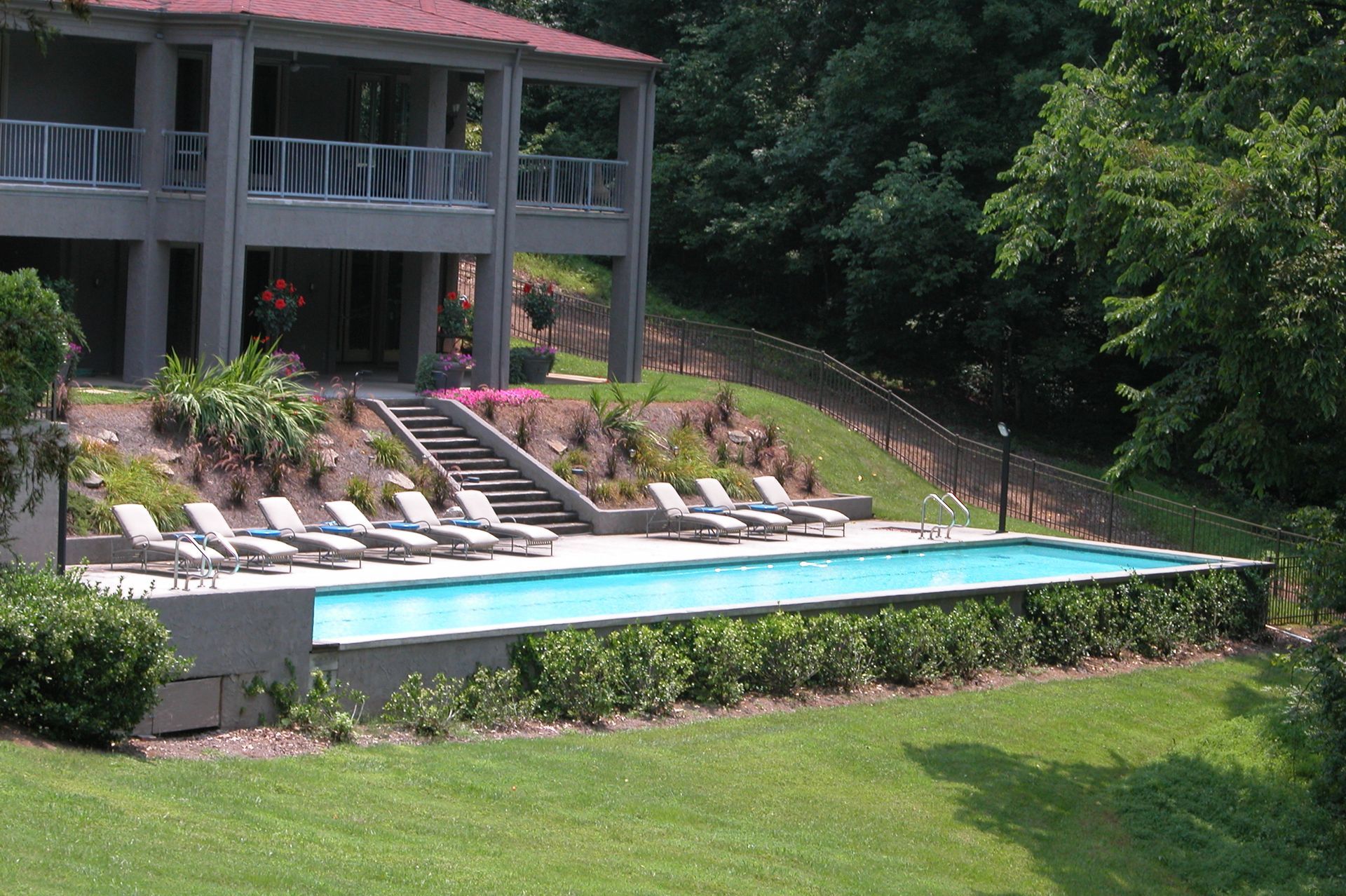 Outdoor pool beside a gray house with red roof, lounge chairs, and lush green lawn