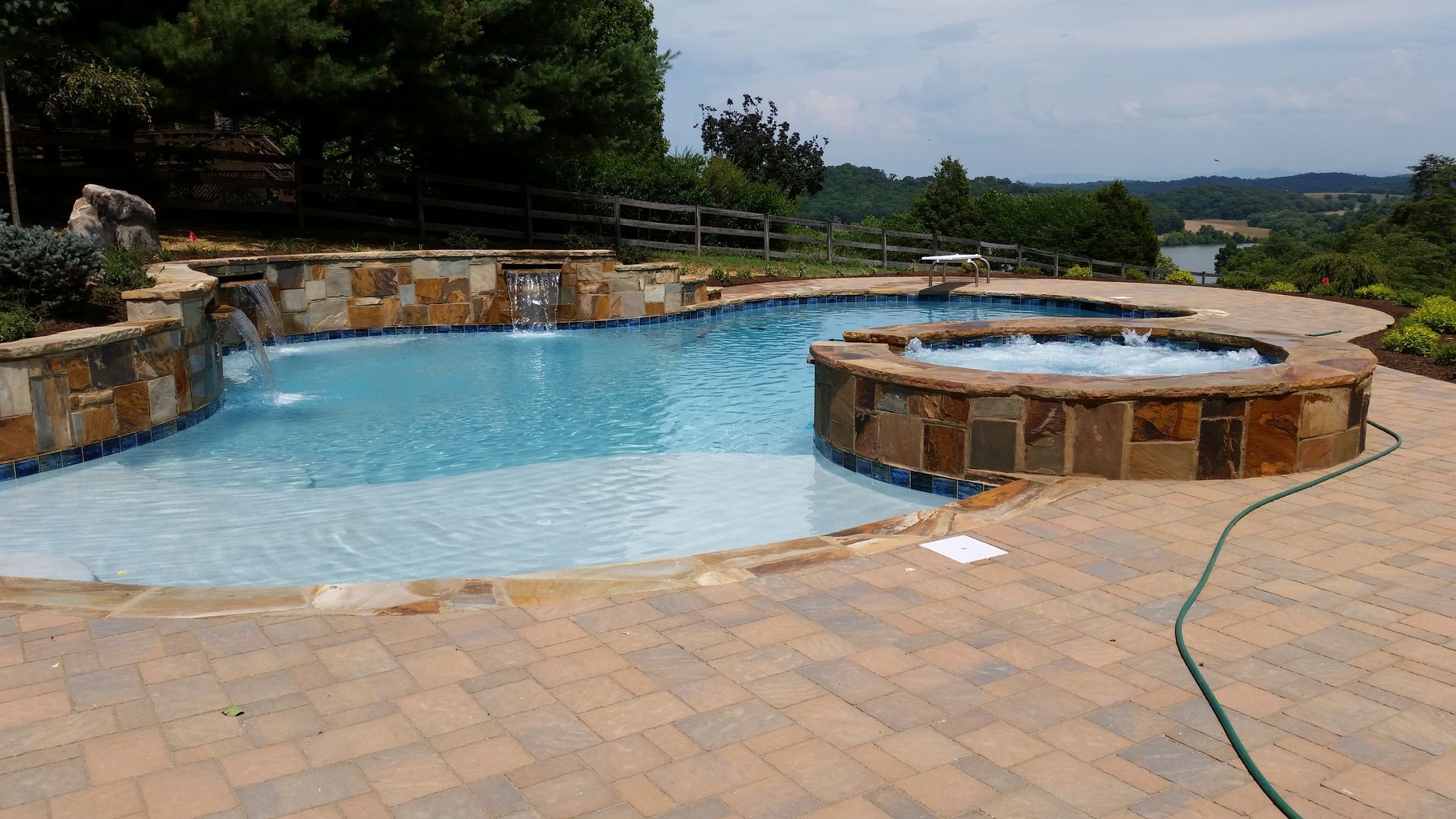 Backyard pool with curved blue water, stone hot tub, and brick patio overlooking trees and hills