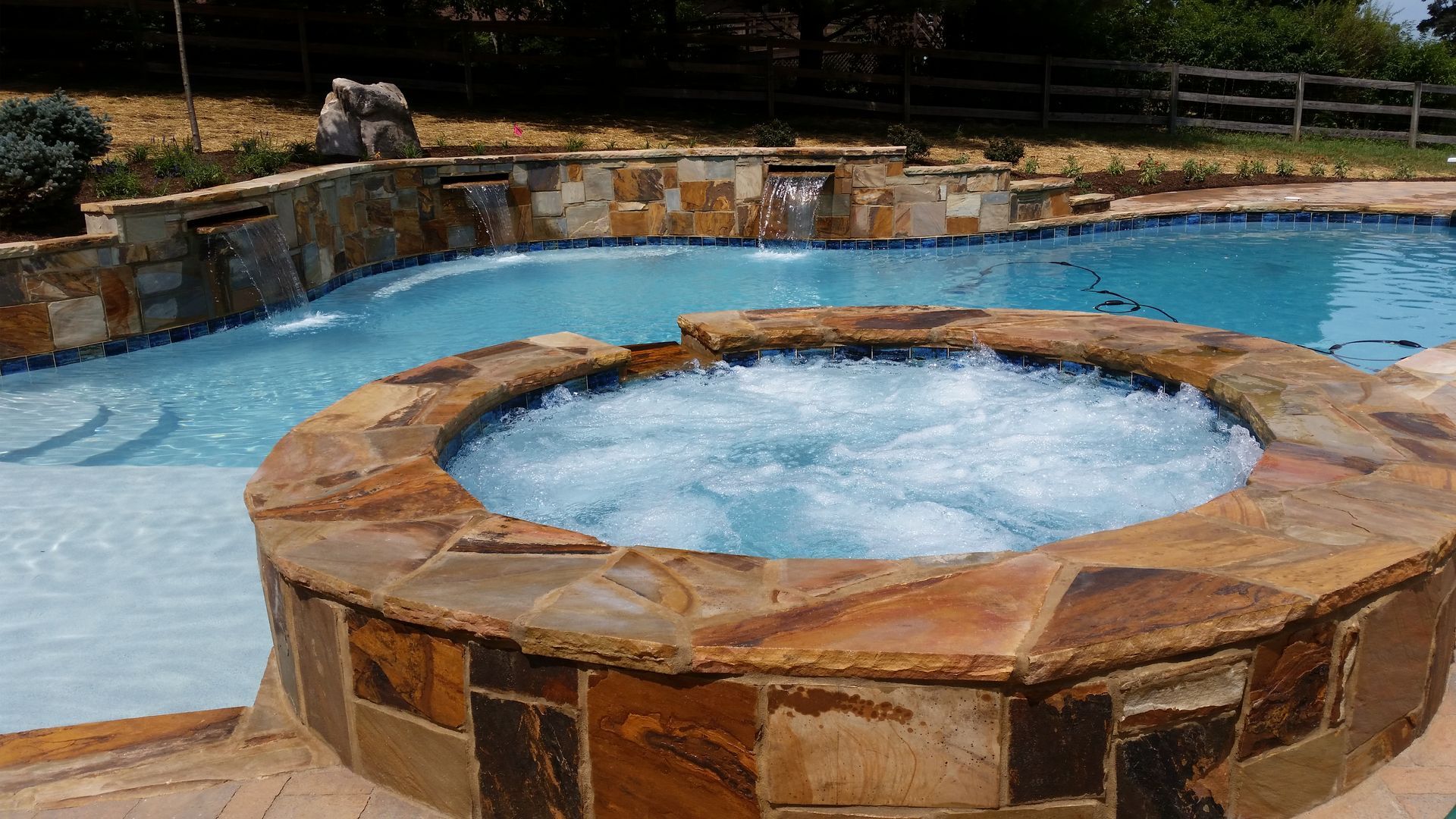 Stone hot tub bubbling in a blue swimming pool, with a stone patio and fence in the background