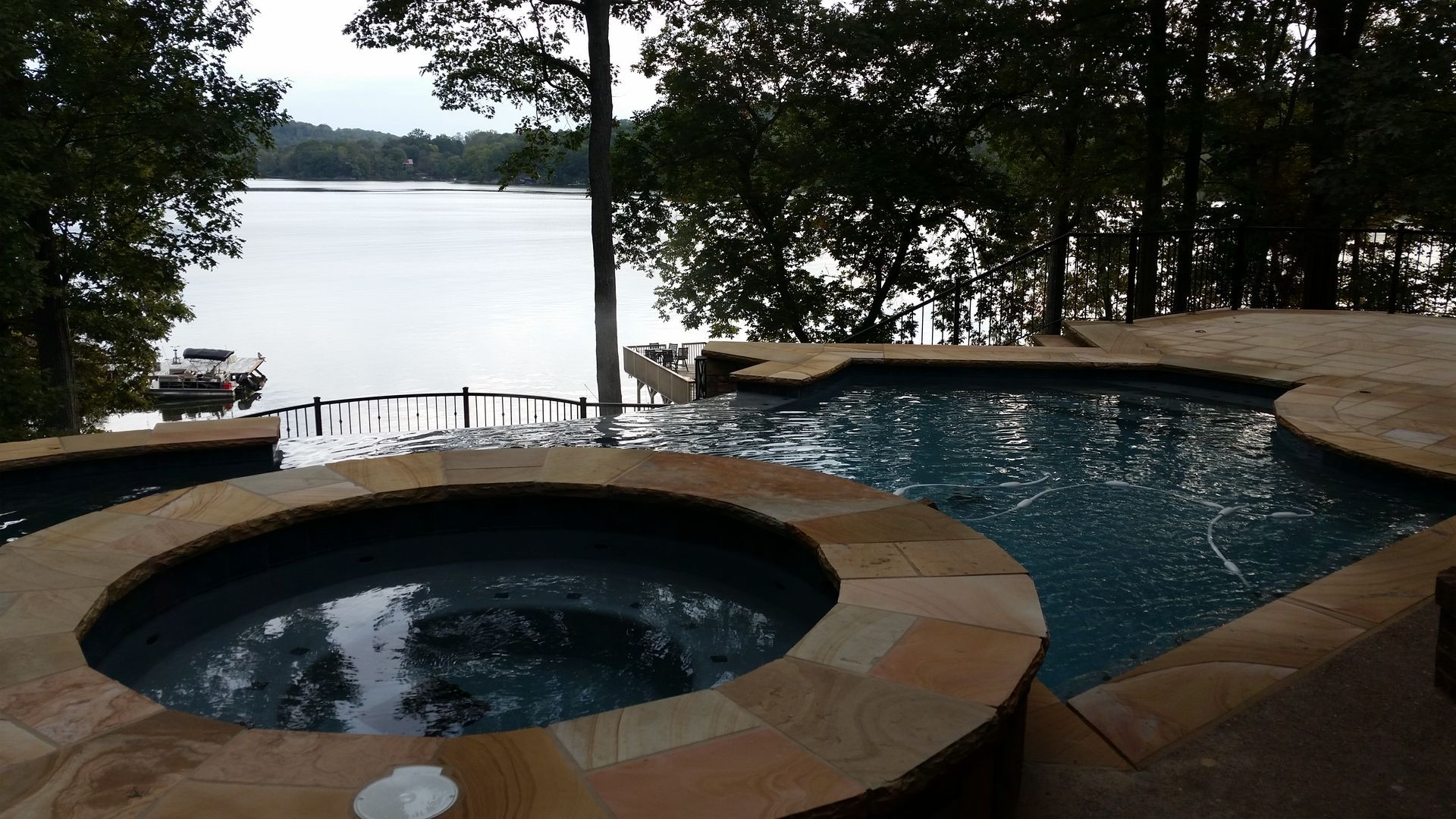 Hot tub and swimming pool overlook a calm lake through trees at dusk