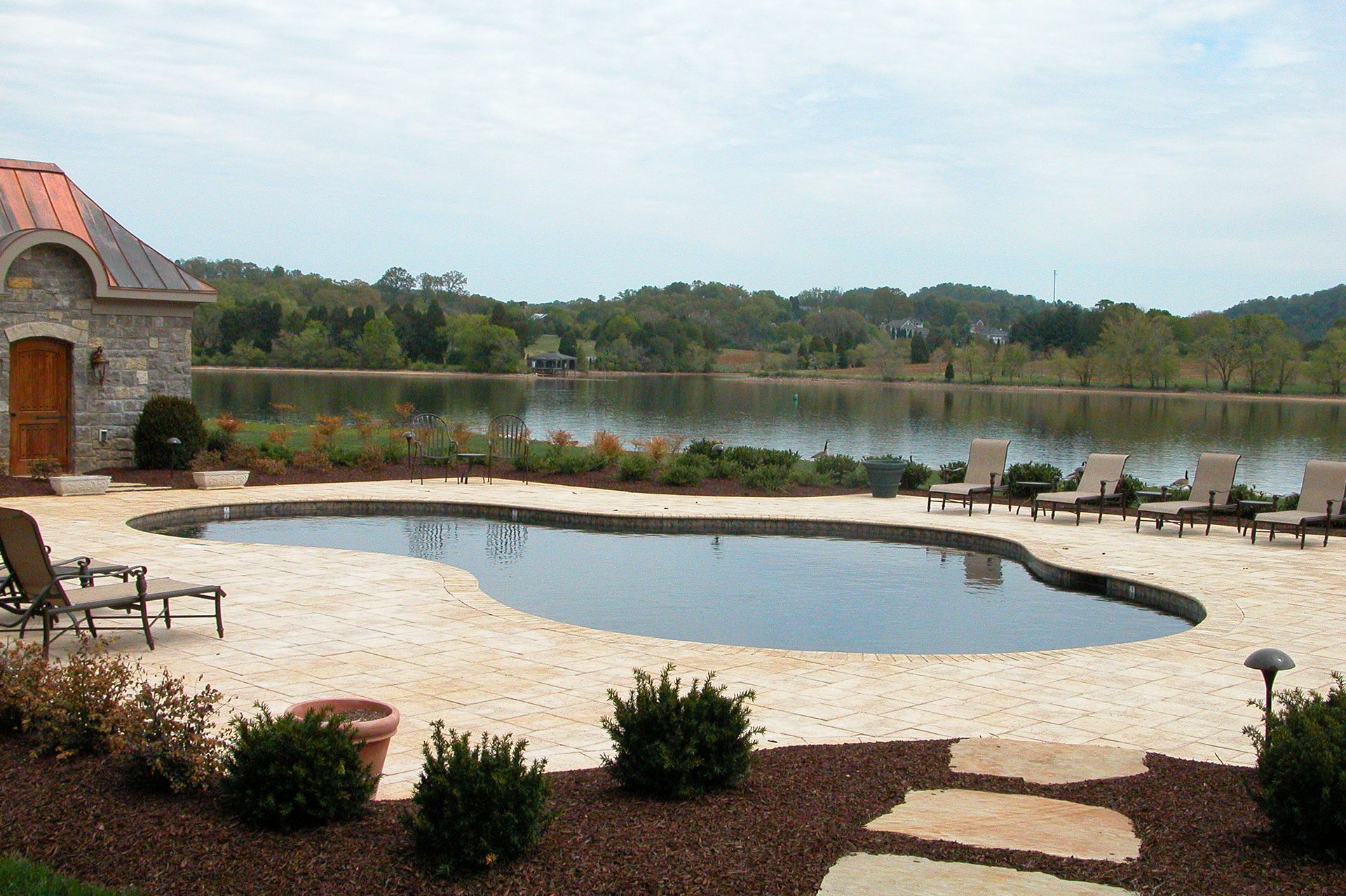 Backyard pool and patio beside a lake, with lounge chairs and a stone building at left.