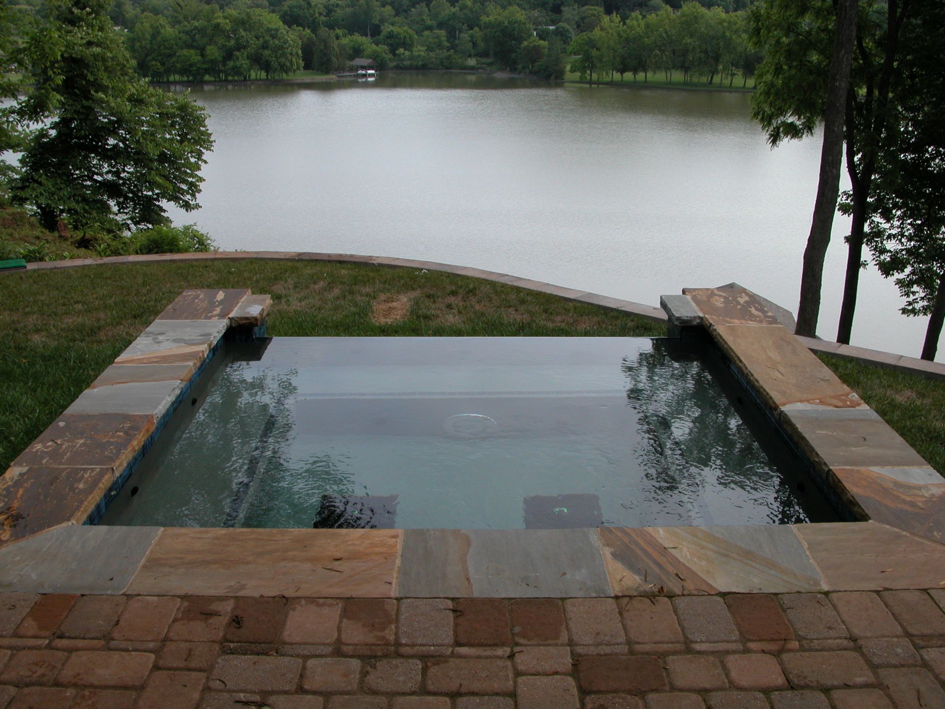Infinity pool overlooking a calm lake, framed by stone patio and trees