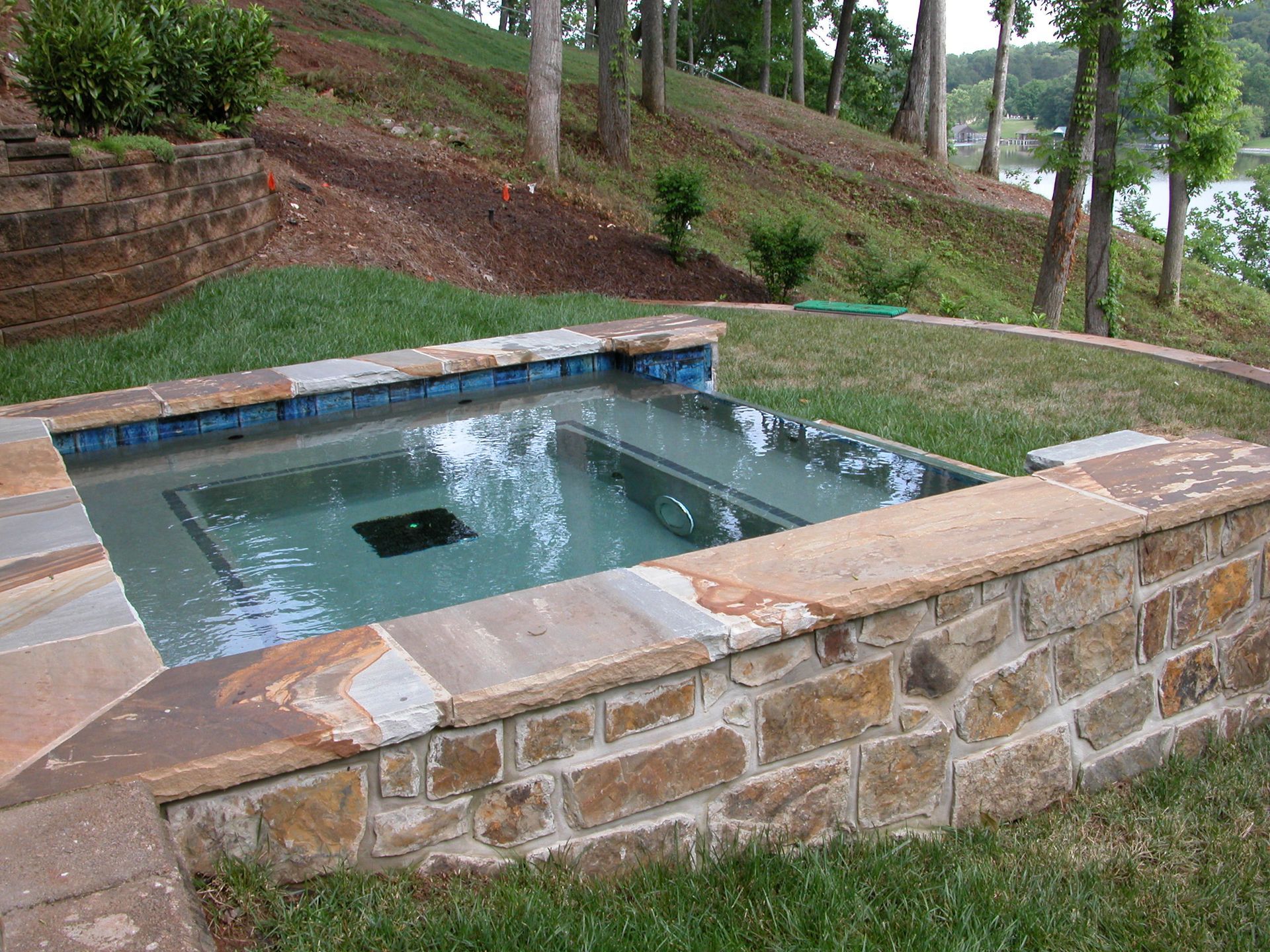 Small outdoor stone-lined pool with green water, set beside a grassy hillside and trees.