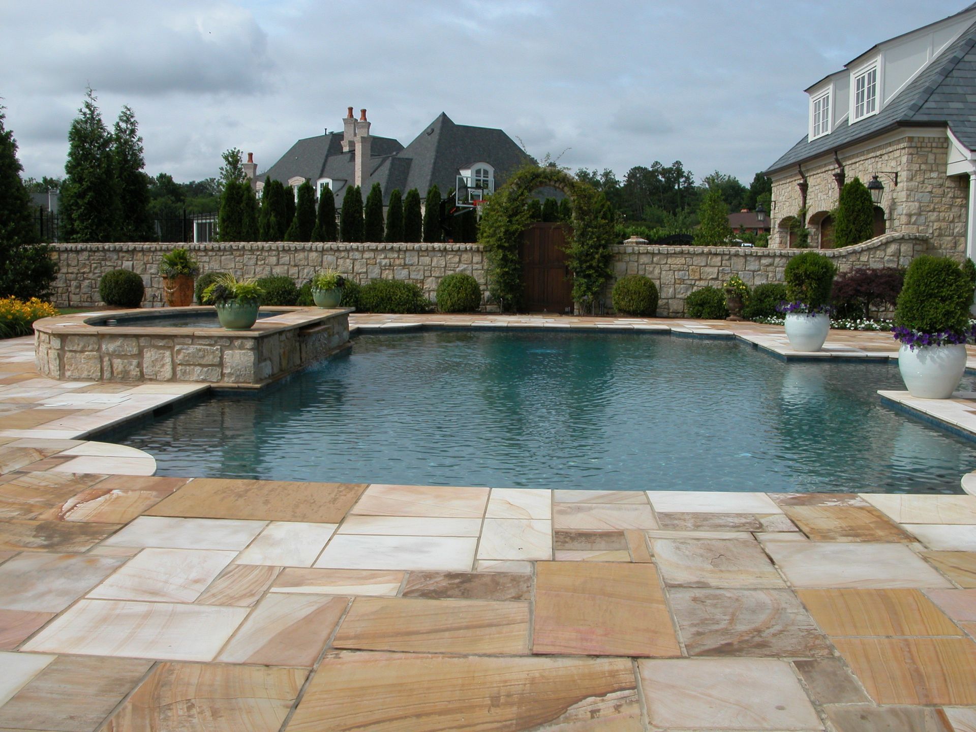 Backyard pool with stone patio, fountains, hedges, and a large house in the background
