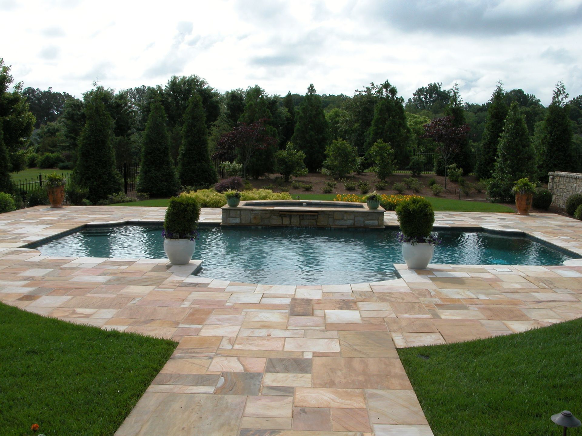 Rectangular backyard pool with stone patio, manicured hedges, and trees under an overcast sky