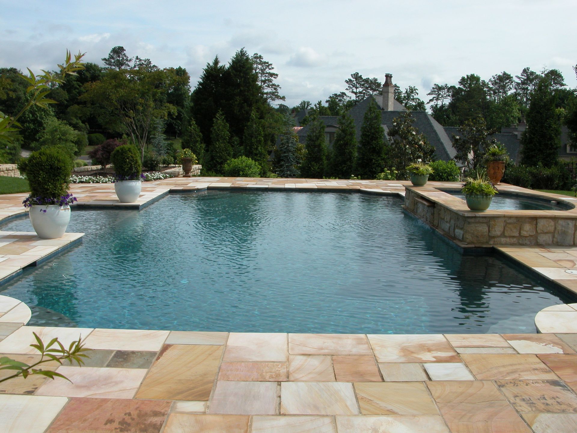 Backyard pool with stone patio, potted plants, and lush trees under a cloudy sky