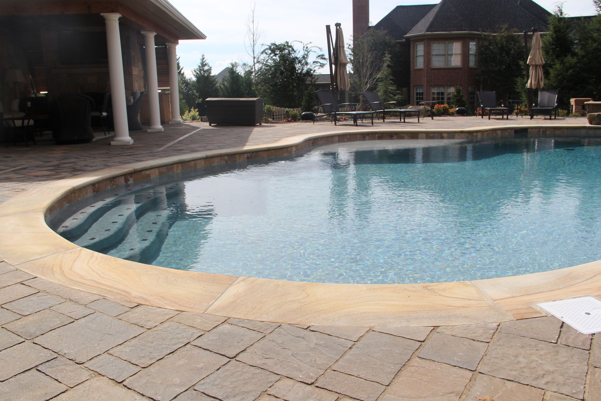 Curved backyard swimming pool with stone patio, patio chairs, and a house in the background
