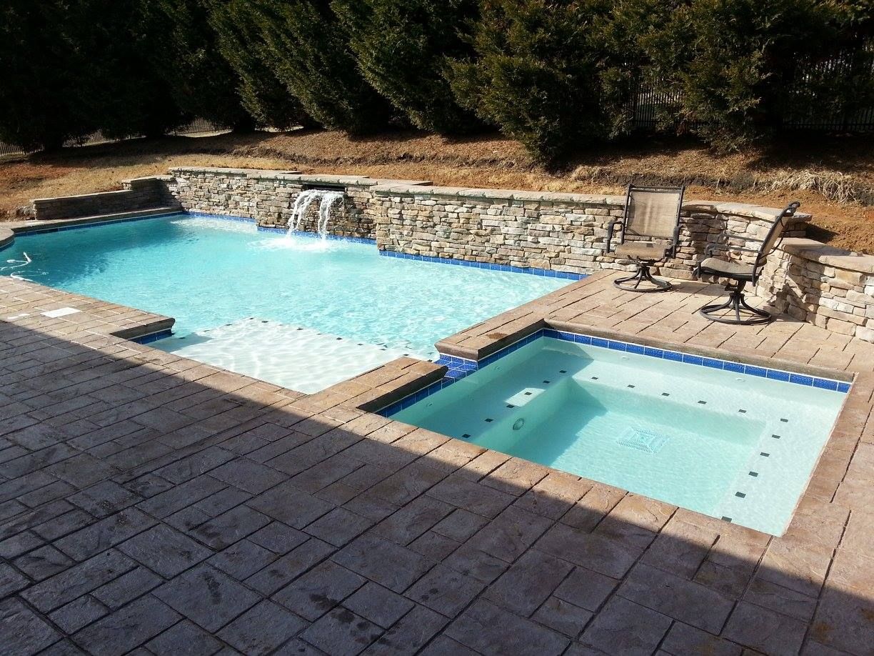 Backyard pool and spa with tan stone deck and lounge chairs, beside a waterfall feature