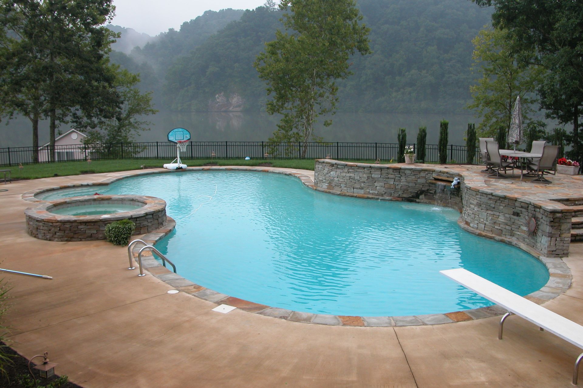 Outdoor swimming pool with a slide, stone wall, and misty lake and trees in the background