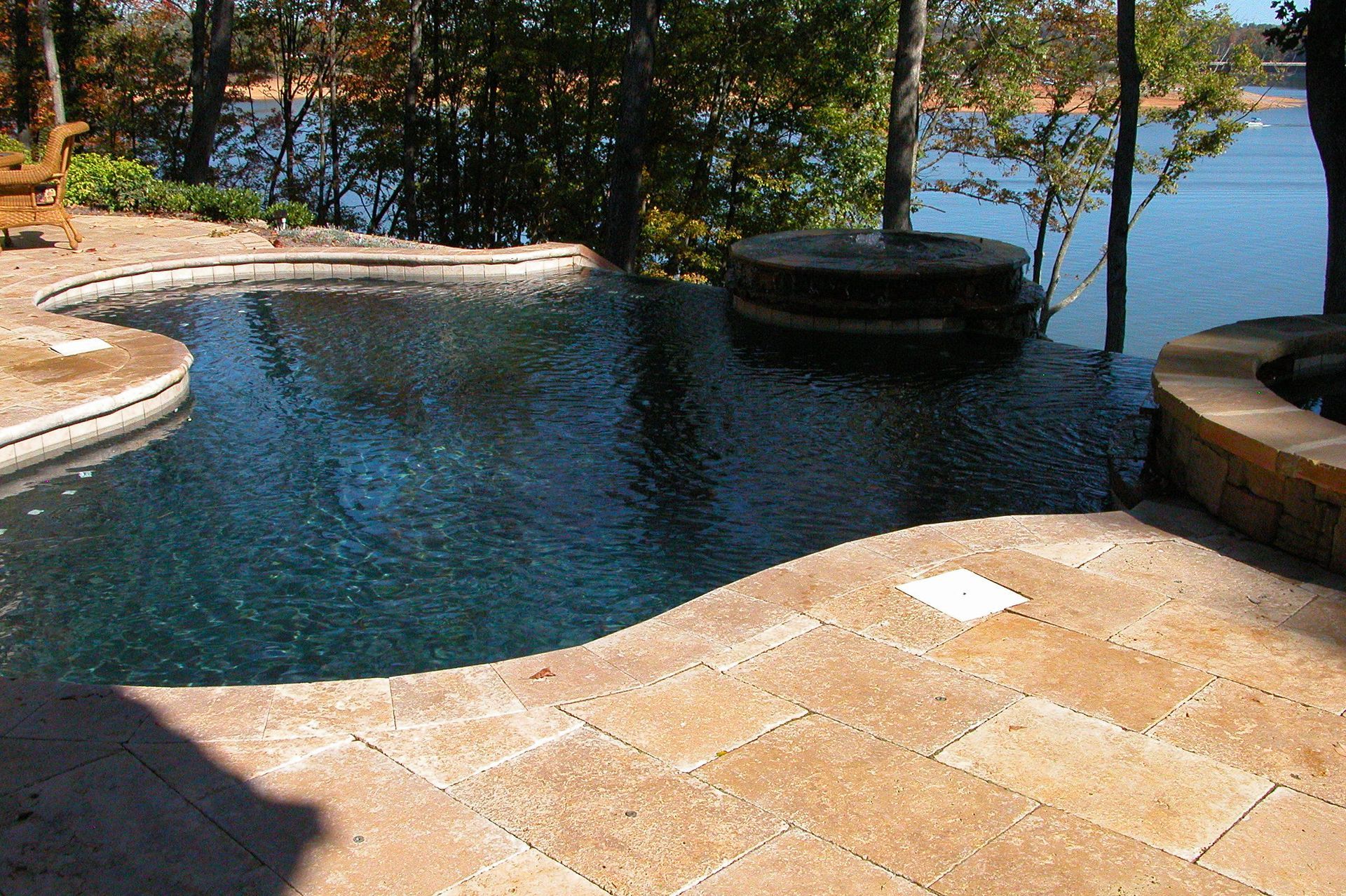 Backyard infinity pool with blue water, tan stone patio, and lake view beyond trees