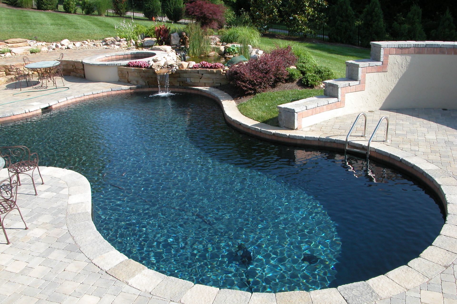Curved backyard swimming pool with stone deck, steps, and landscaped garden